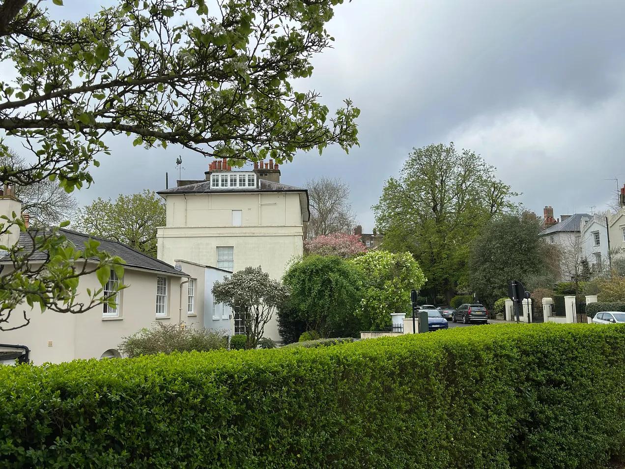 Historic cream-colored townhouse with brick chimneys behind manicured green hedges and spring foliage.