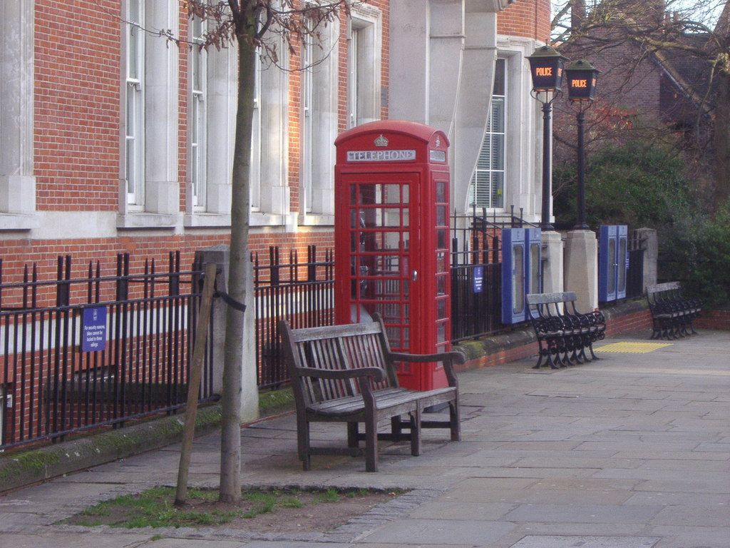 Red British telephone box on a paved street with benches, brick buildings, and a police box in the background.