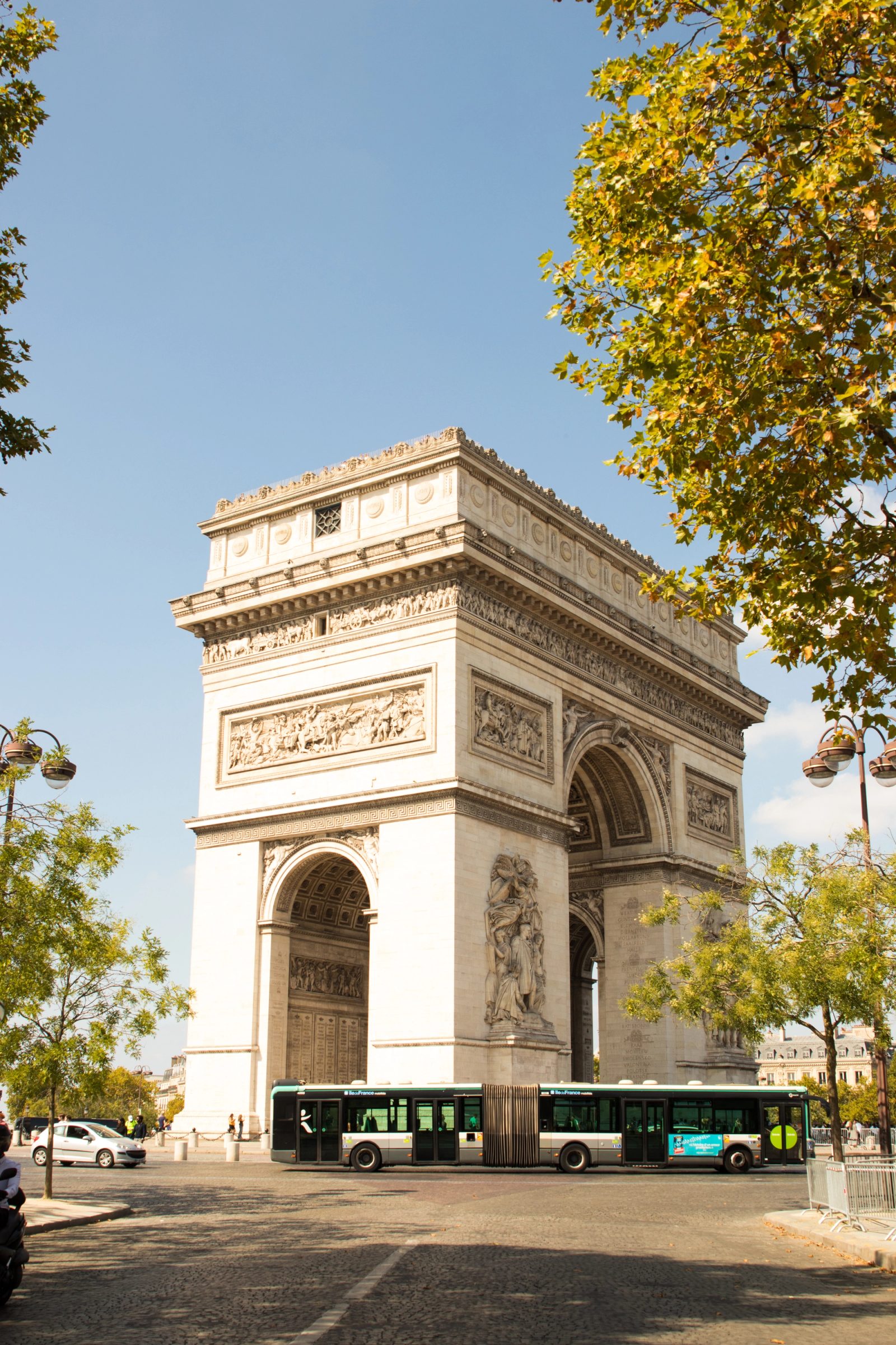 The Arc de Triomphe in Paris photographed from ground level with yellow autumn trees framing both sides and a green public bus in the foreground.