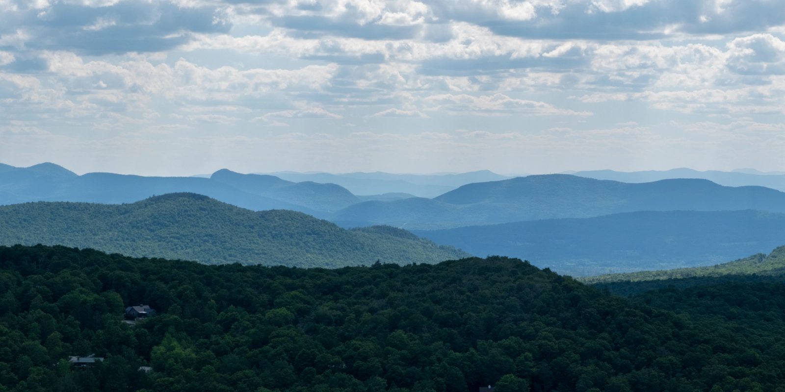 A landscape photograph showing multiple layers of forested mountain ridges receding into the distance under a cloudy sky, with dark green vegetation in the foreground and blue-toned mountains fading into the hazy horizon.