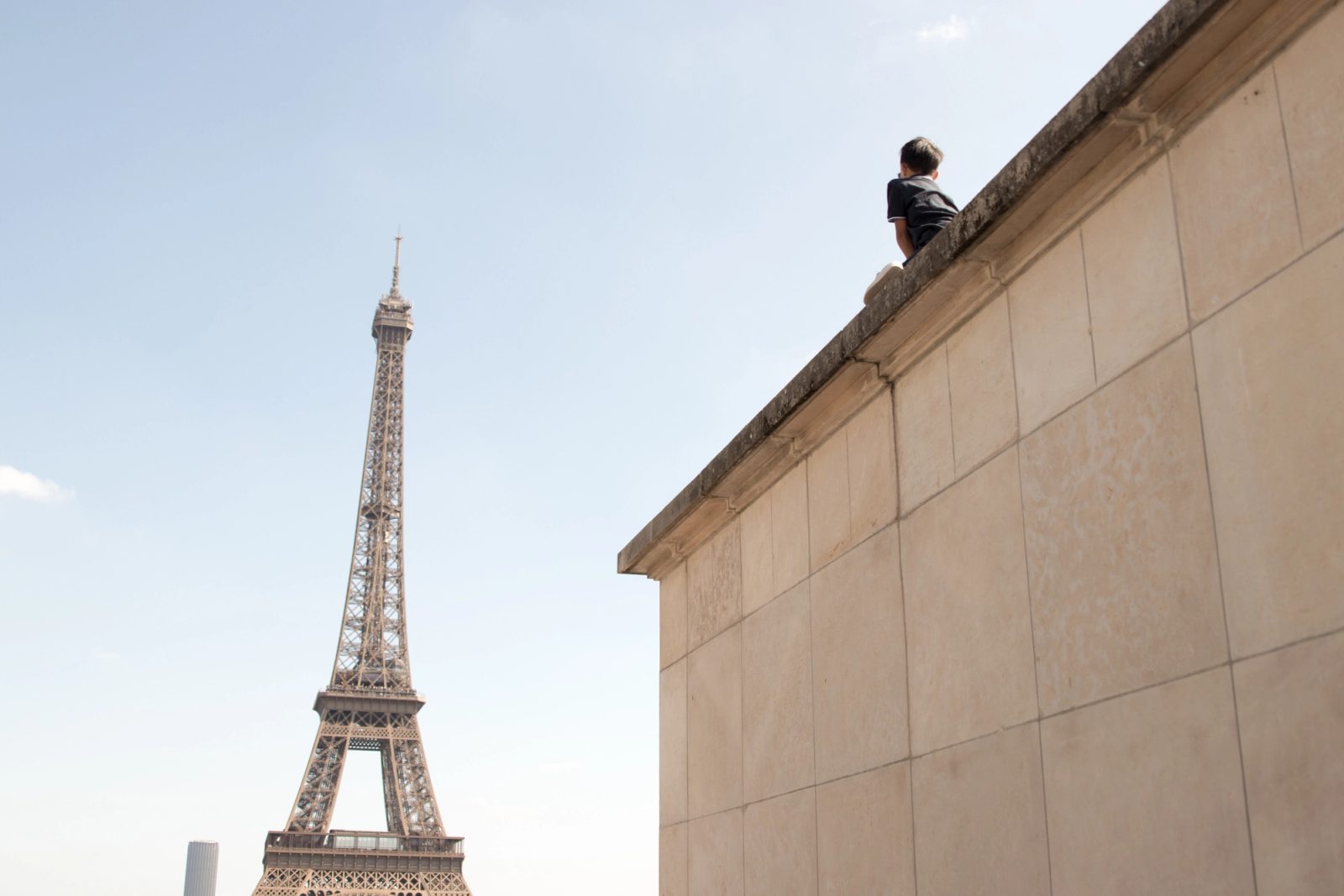 A boy sits on the upper edge of a beige stone building looking toward the Eiffel Tower in Paris under a pale sky.