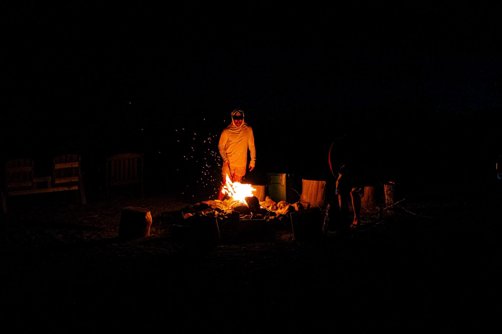 A person wearing an orange blanket or robe stands beside a crackling campfire at night, surrounded by wooden stumps and complete darkness.