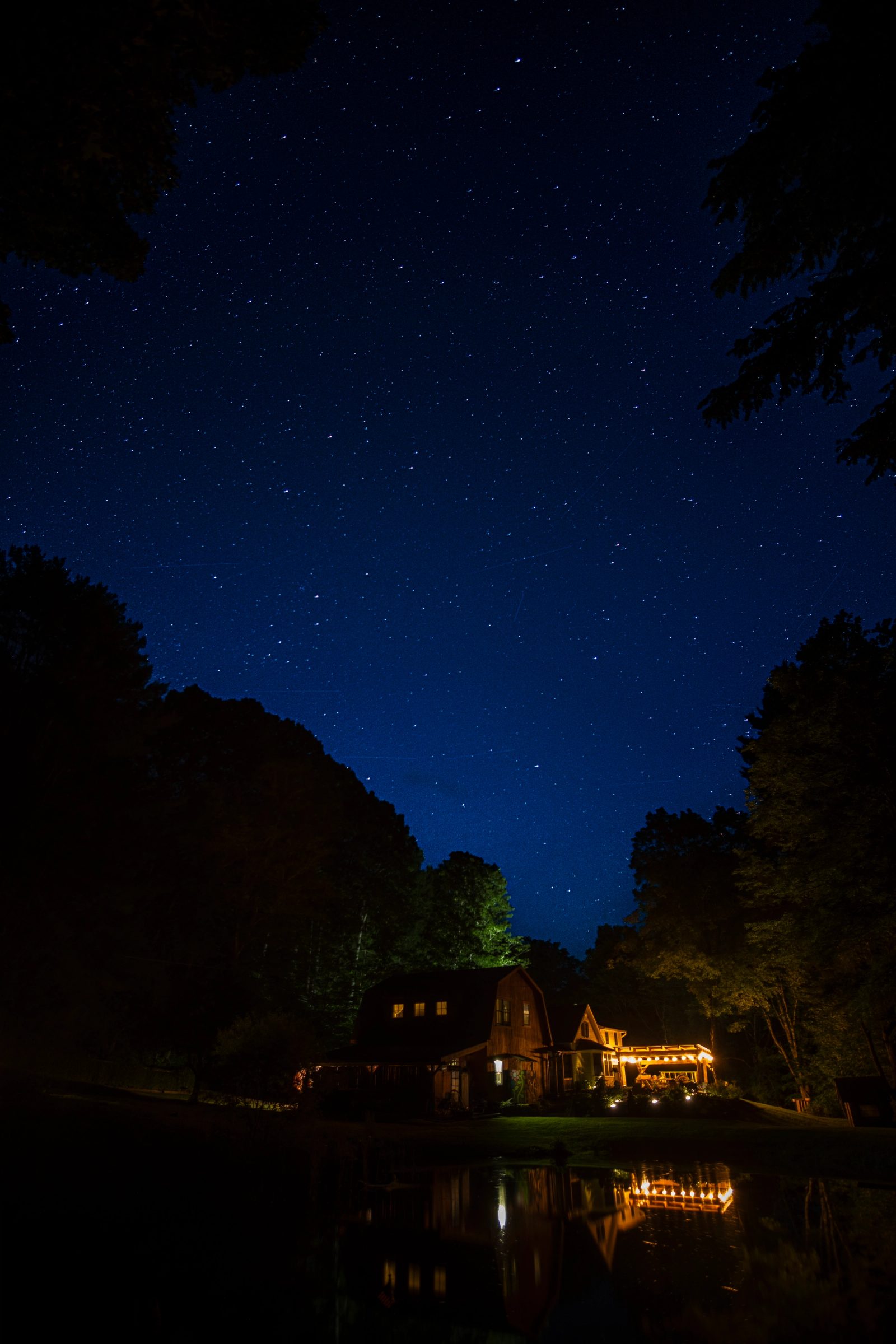 A wooden house with illuminated windows sits beside a pond at night, surrounded by trees with a starry deep blue sky overhead and reflections in the water.