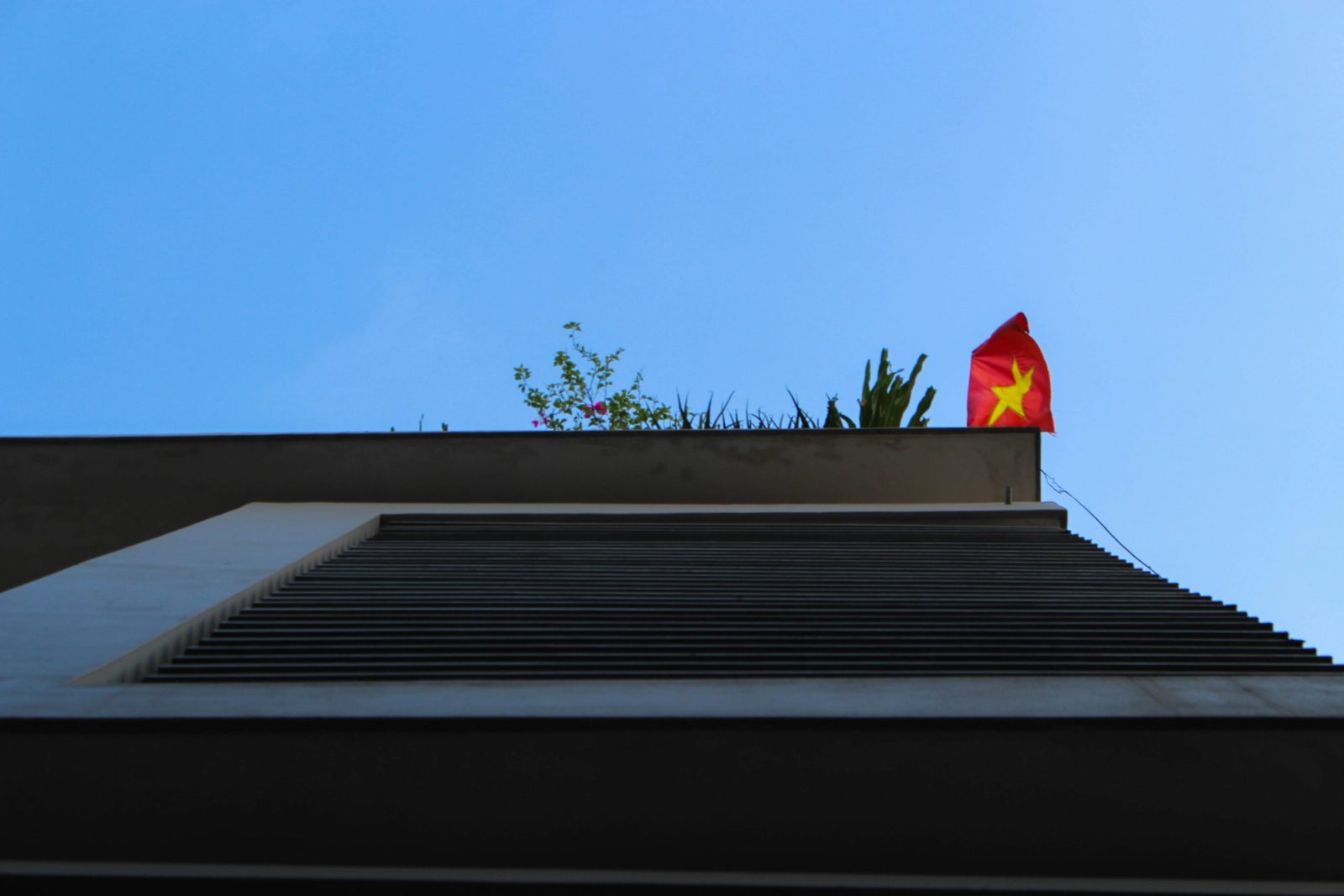 A red flag with a yellow star flies on top of a modern concrete building with horizontal slats, photographed from below against a clear blue sky.