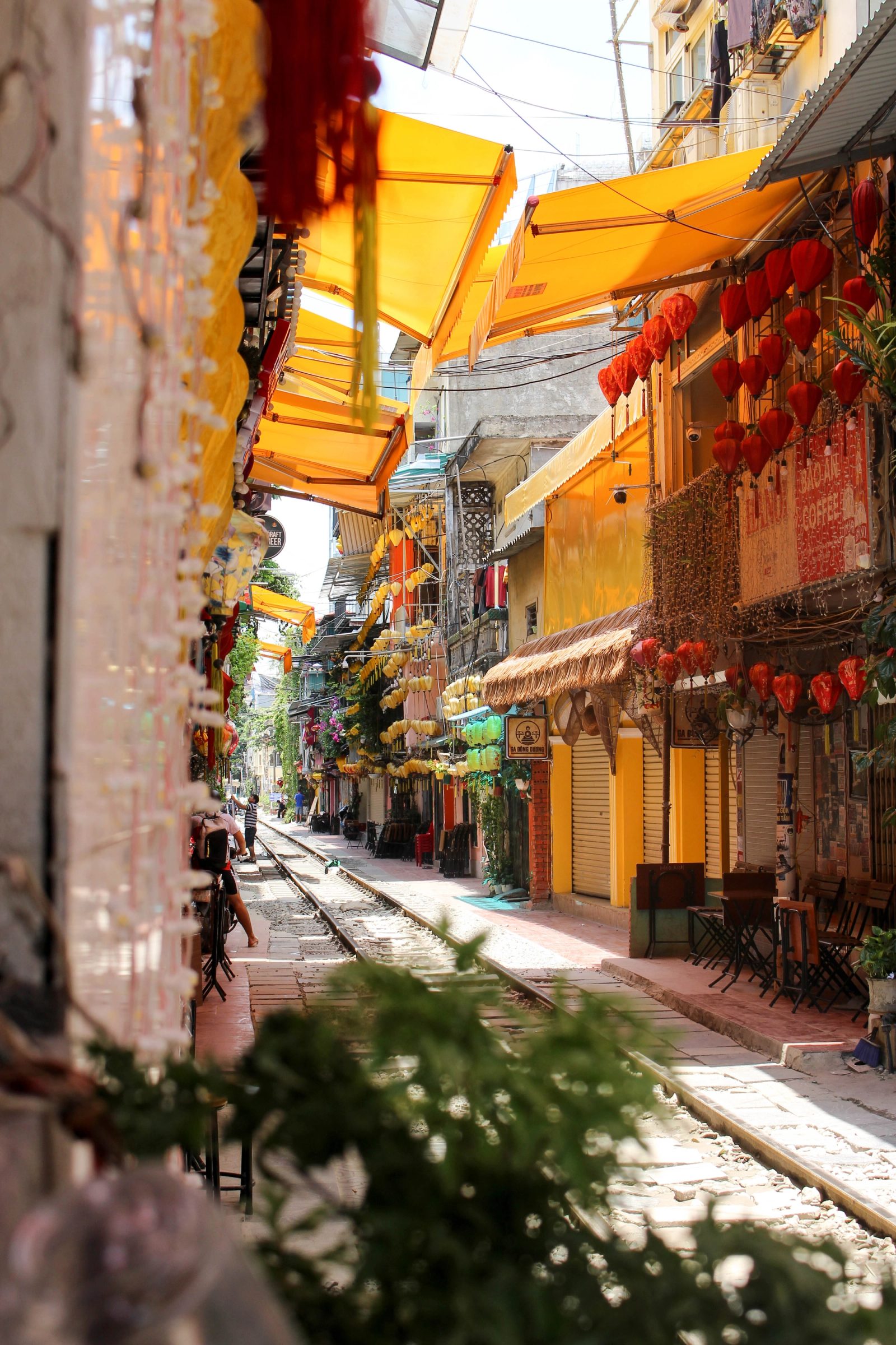 A narrow alley with railway tracks running through the center, lined with residential buildings, yellow sun awnings overhead, red lanterns hanging from facades, and green foliage in the foreground.
