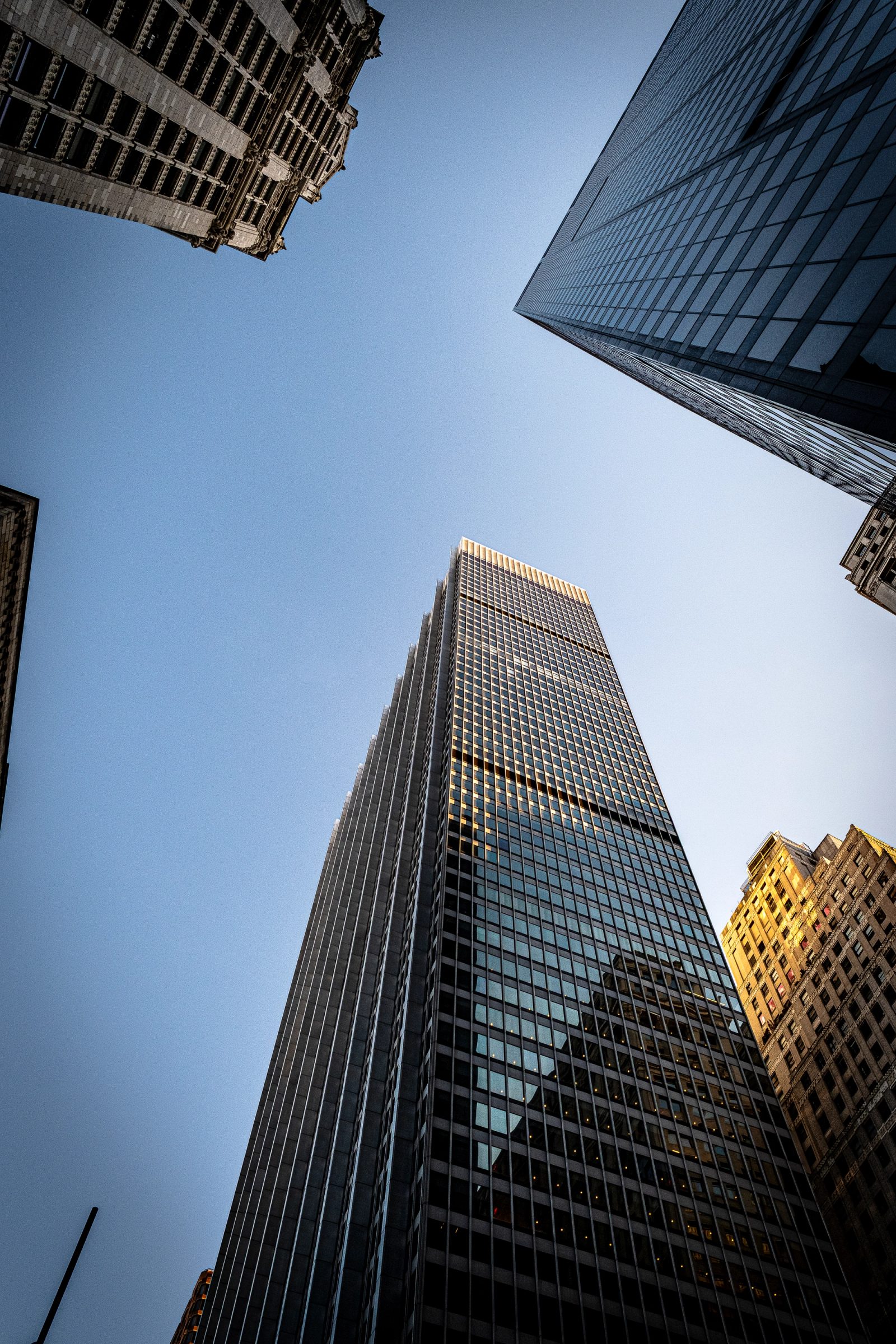A low-angle photograph looking up at multiple tall skyscrapers converging toward the sky, with glass and steel facades in shadow and golden light against a clear blue sky.