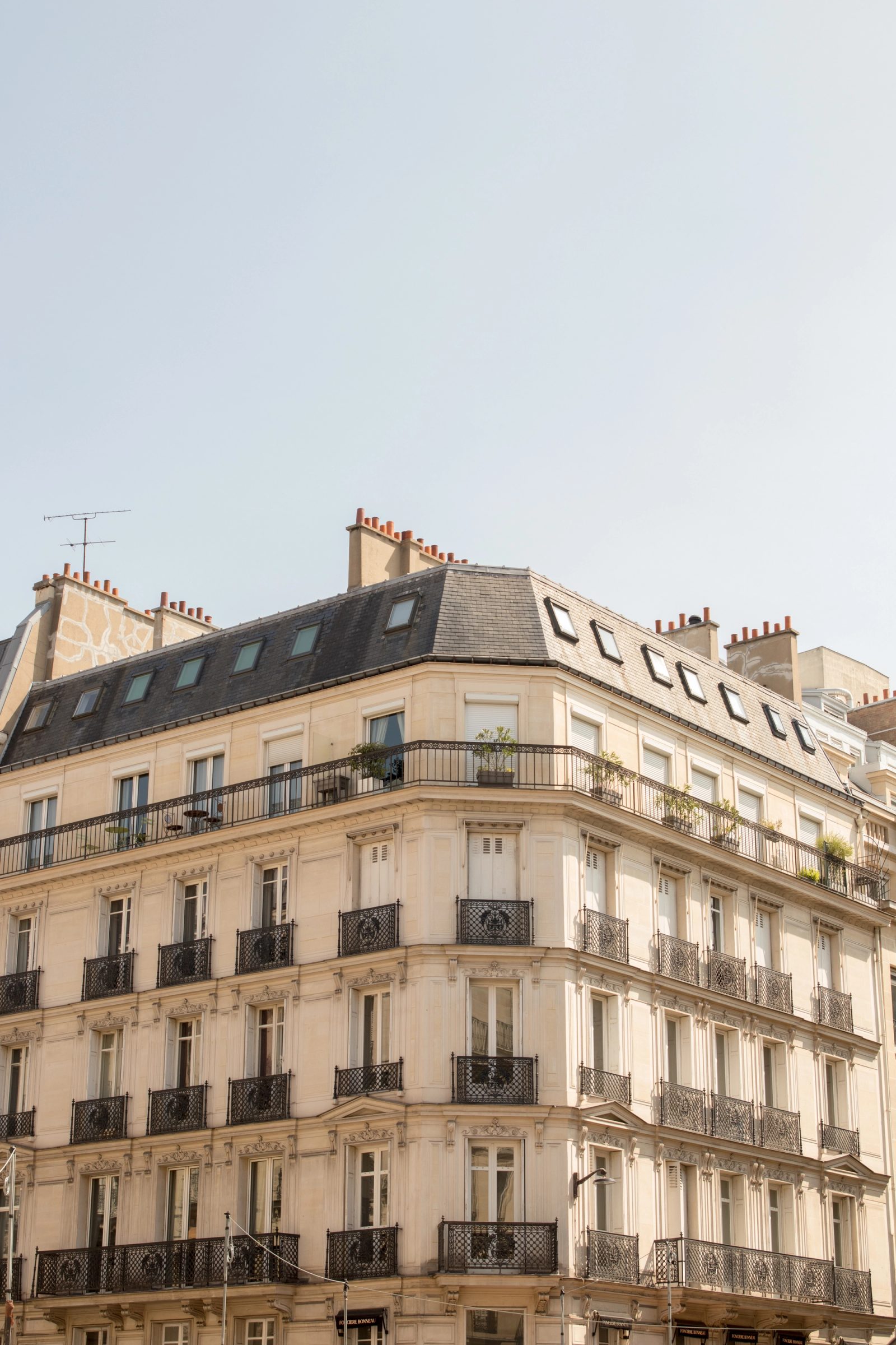 A classic Parisian apartment building with cream stone facade, wrought iron balconies, mansard roof with dormer windows, and terracotta chimney pots against an overcast sky.