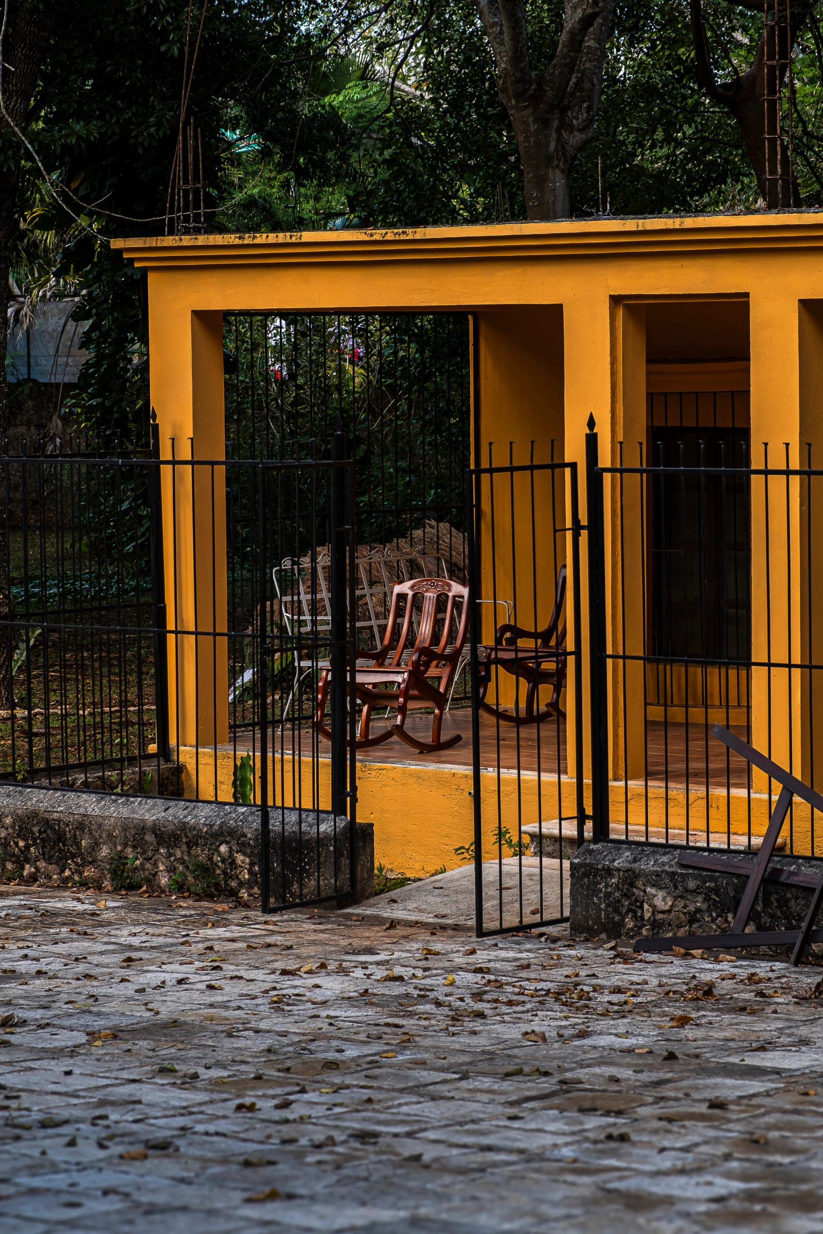 A bright yellow open-air pavilion with terracotta floor contains striped rocking chairs behind a black iron gate, backed by dense tropical trees.