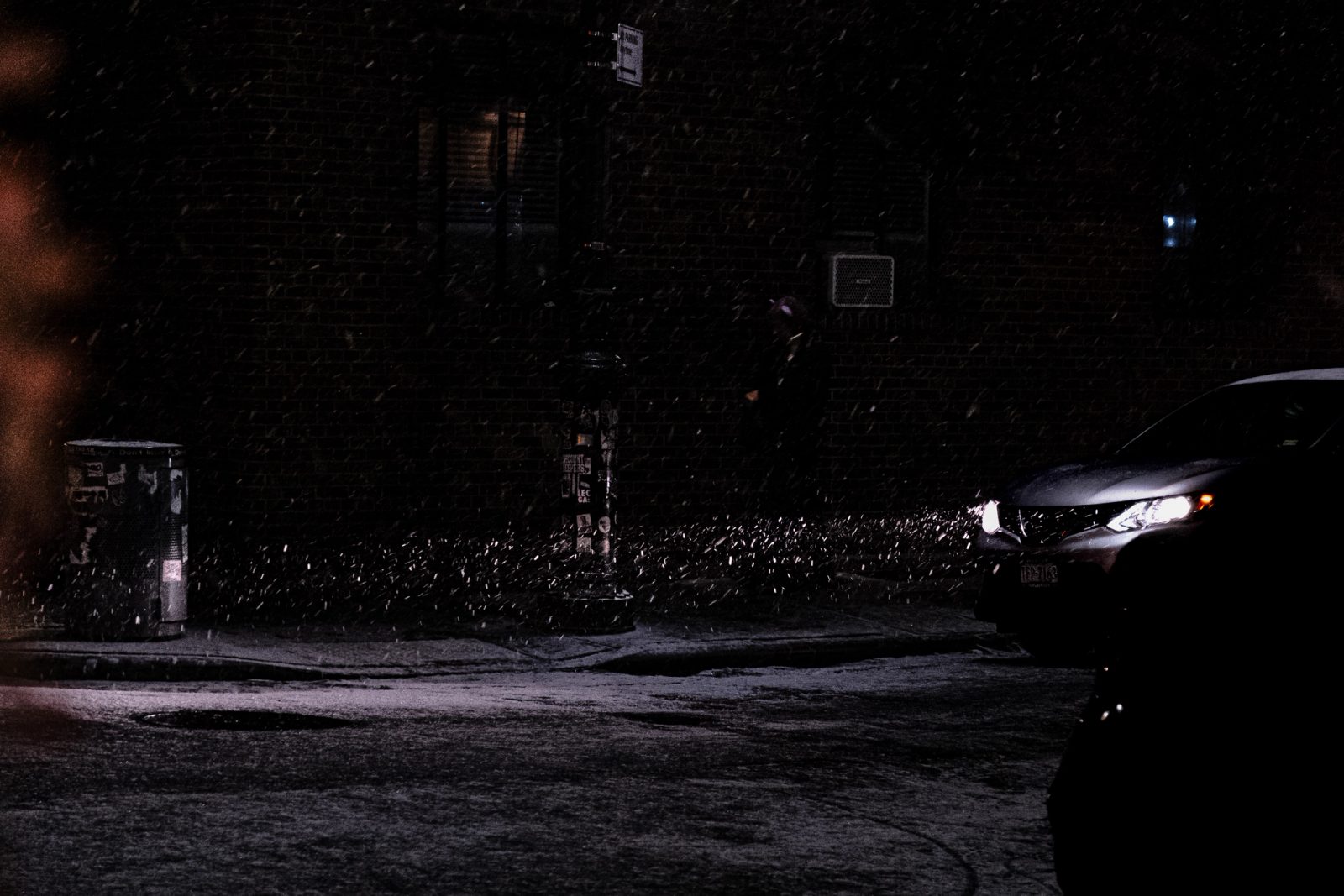 A dark sedan with illuminated headlights stopped at a snowy nighttime intersection with traffic lights and street signs visible above.