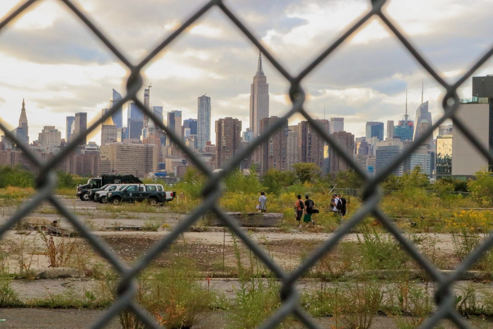 View of a city skyline with the Empire State Building visible through a chain-link fence, with people and vehicles in an overgrown vacant lot in the foreground.