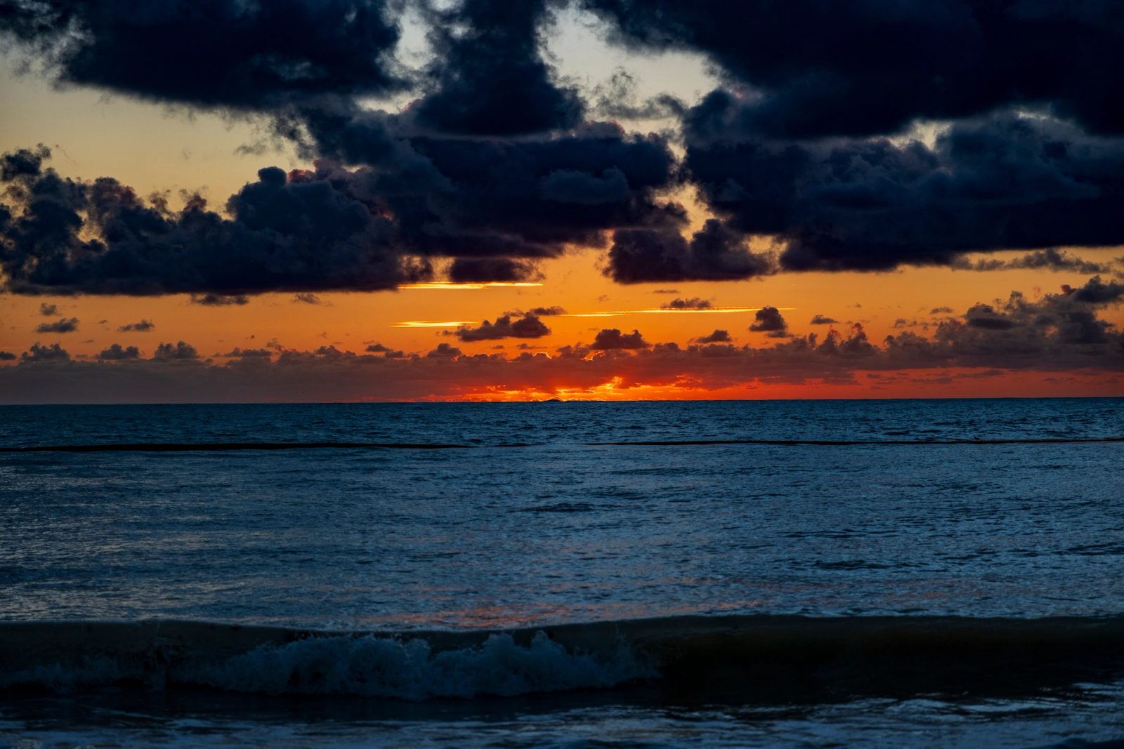 A dramatic ocean sunset with brilliant orange and yellow light breaking through heavy dark storm clouds above dark blue waves.