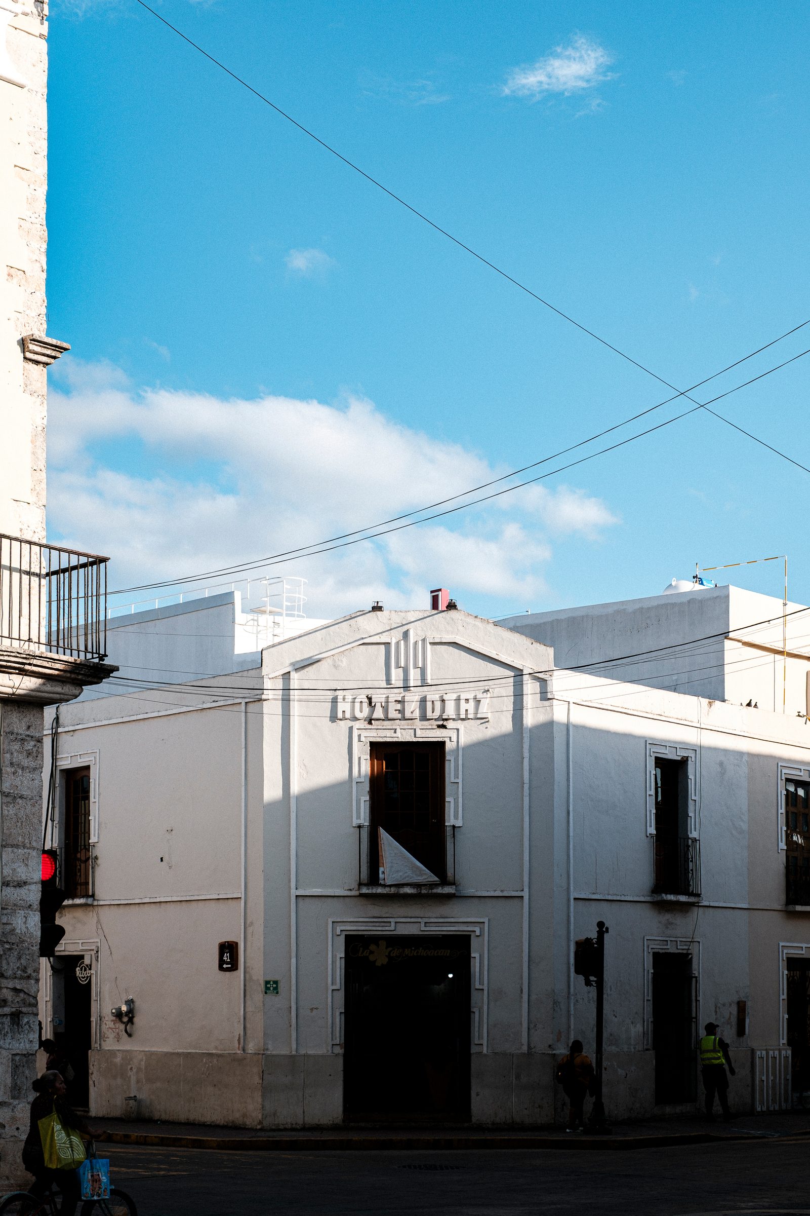 A white corner building with 'Hotel Dirt' text on its facade, with dark open windows and doorways, pedestrians walking in shadow, and power lines crossing a blue sky overhead.