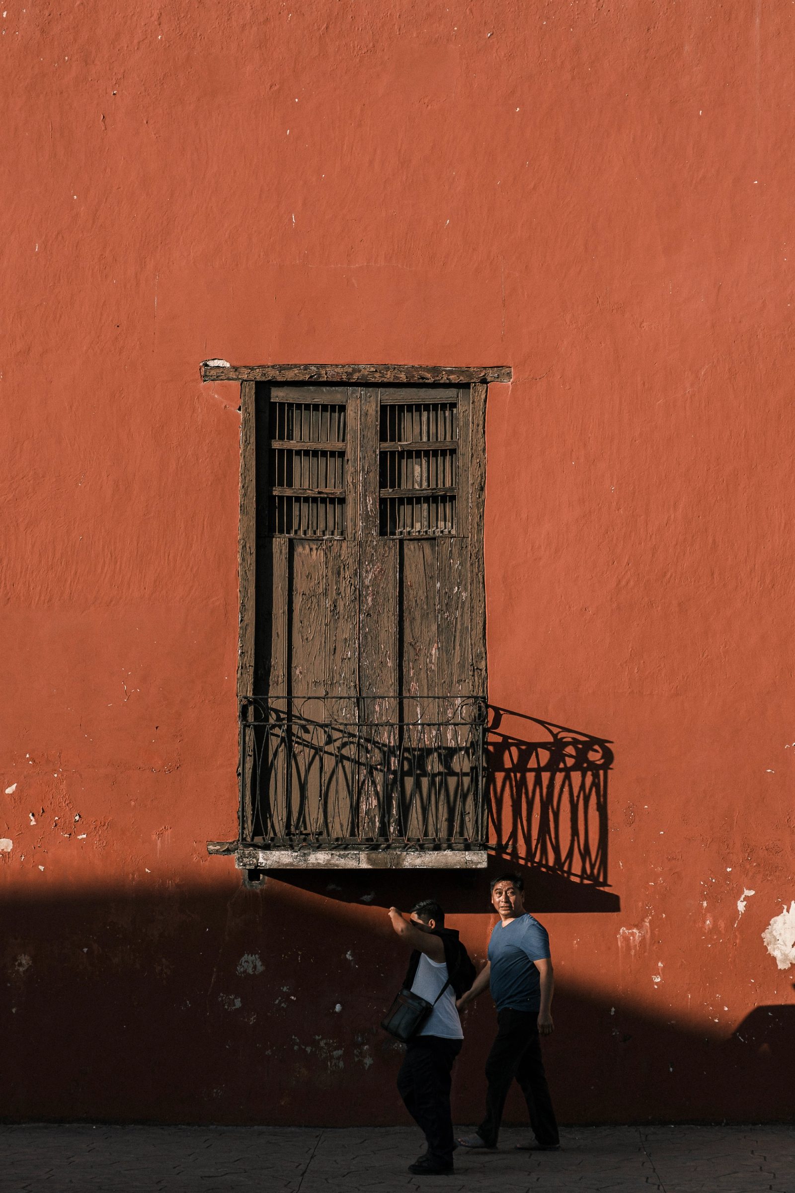 Two people walking beneath a weathered wooden balcony door with decorative ironwork against a large terracotta-colored wall.