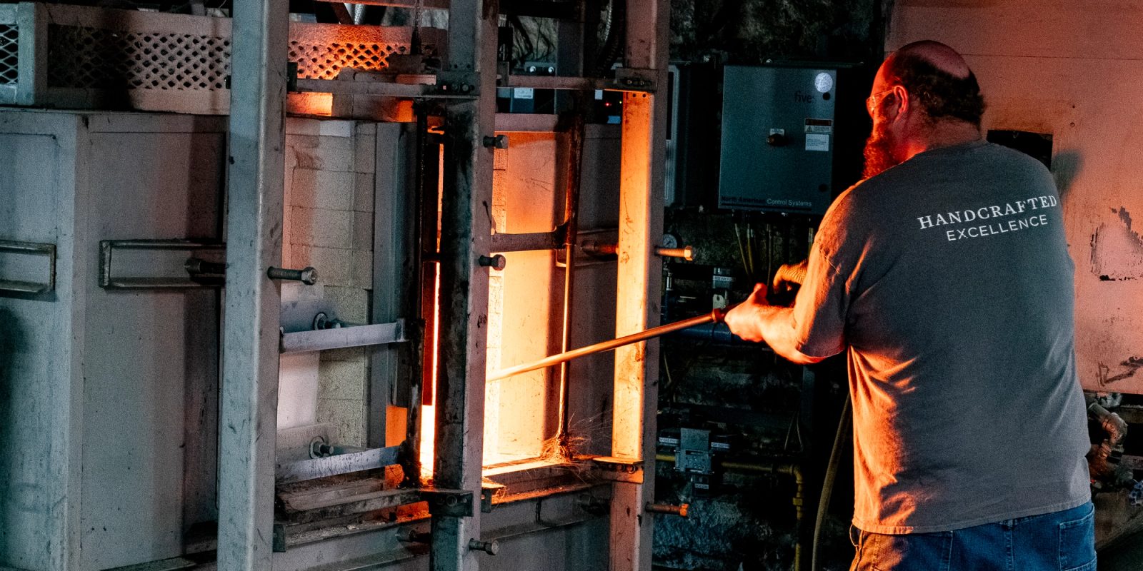 A glassblower in a vest labeled 'Handcrafted Excellence' works at an open industrial furnace, holding a long rod toward the glowing orange heat.