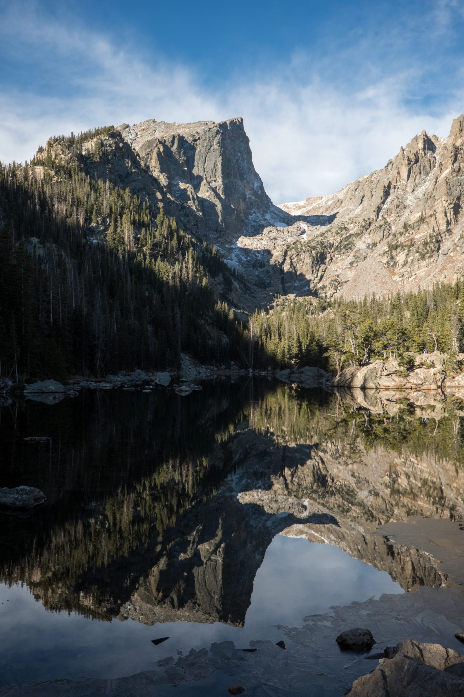 A dramatic rocky mountain peak with forested slopes reflects perfectly in a calm alpine lake under blue sky with wispy clouds.