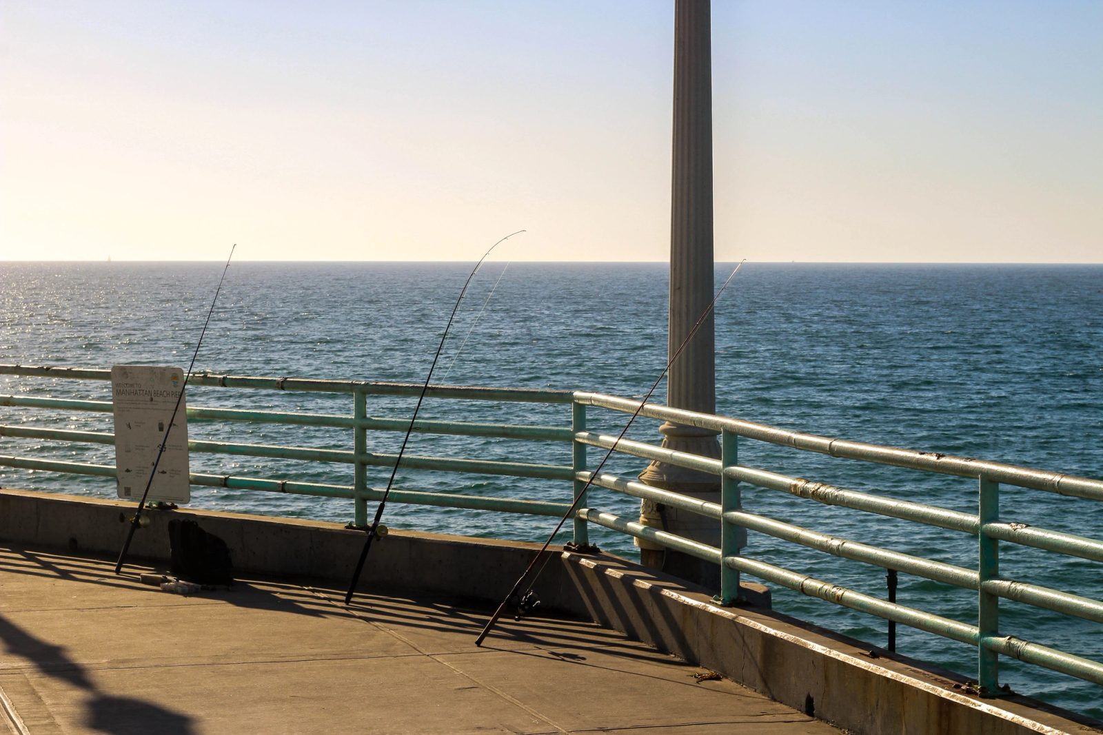 Two fishing rods positioned on a concrete pier deck with metal railings overlooking a calm ocean under a clear sky.