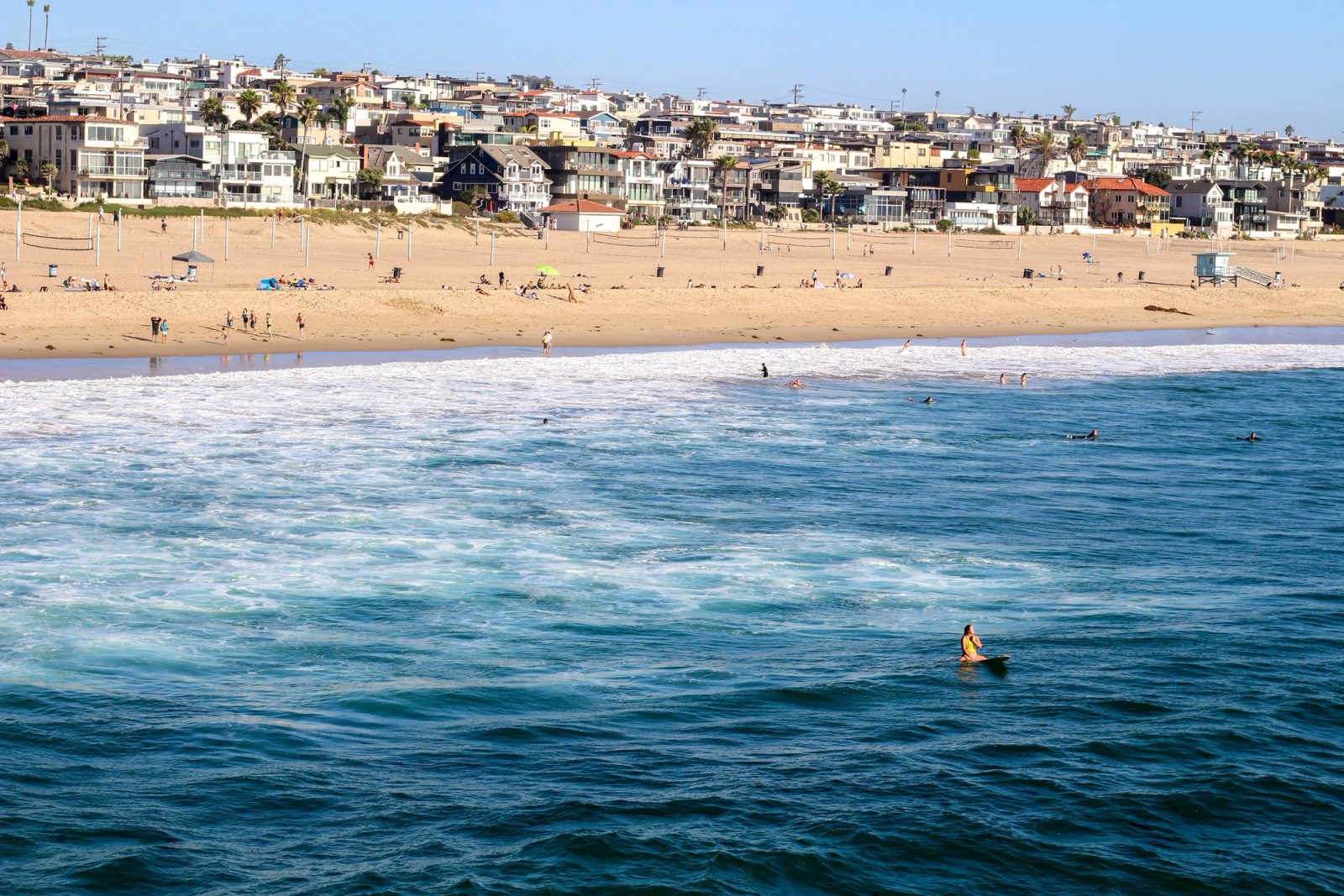 A surfer in a yellow shirt sits on their board in blue-green ocean water with a sandy beach and residential buildings visible in the background.