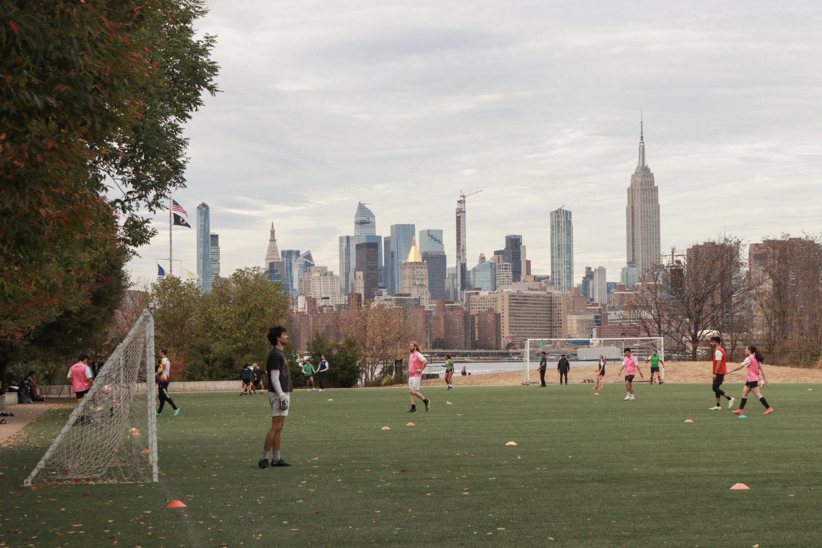 Soccer players on a field with the Empire State Building and Manhattan skyline visible in the background under an overcast sky.