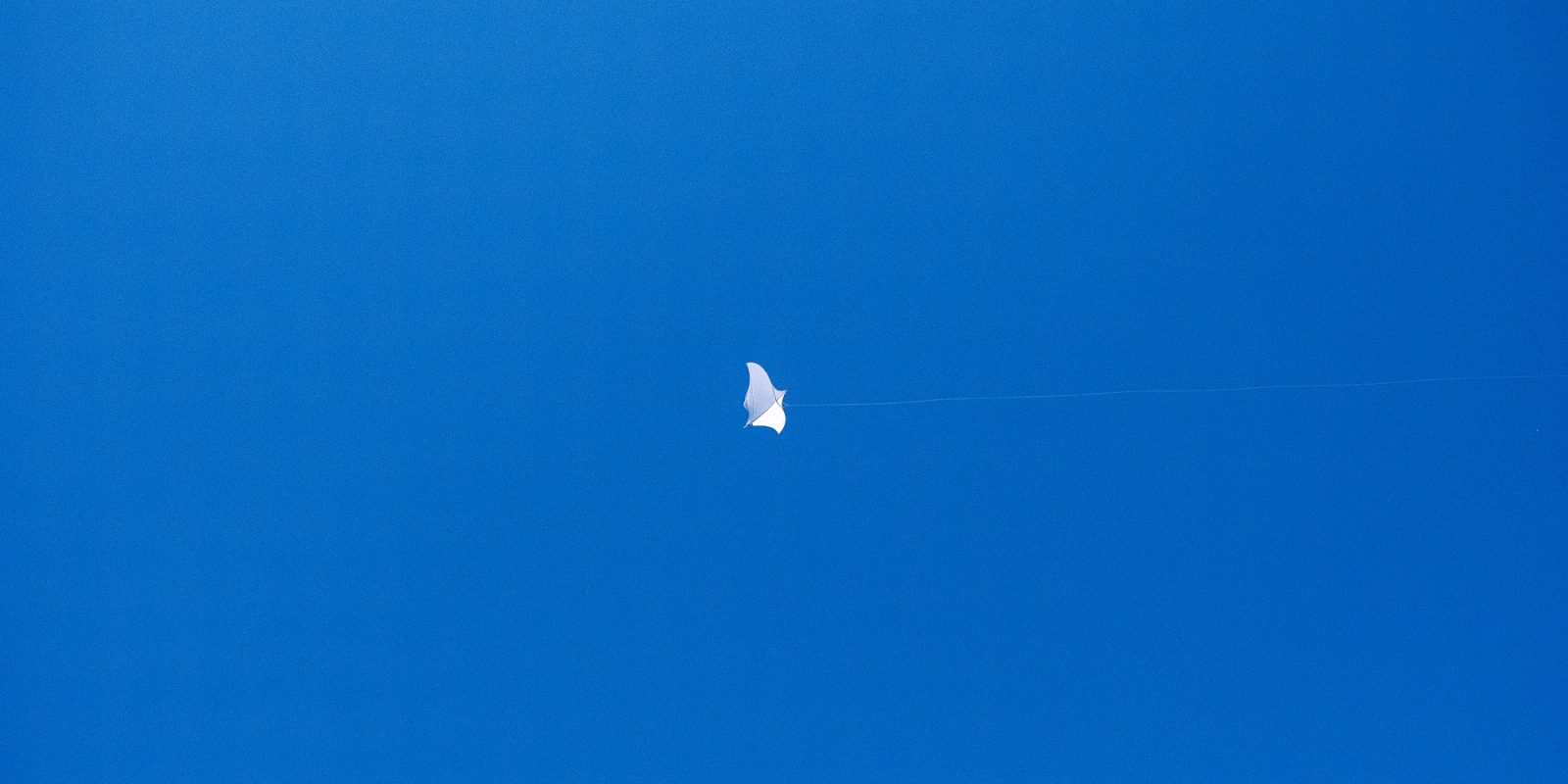 A white diamond-shaped kite with a thin line attached flies against a clear blue sky.