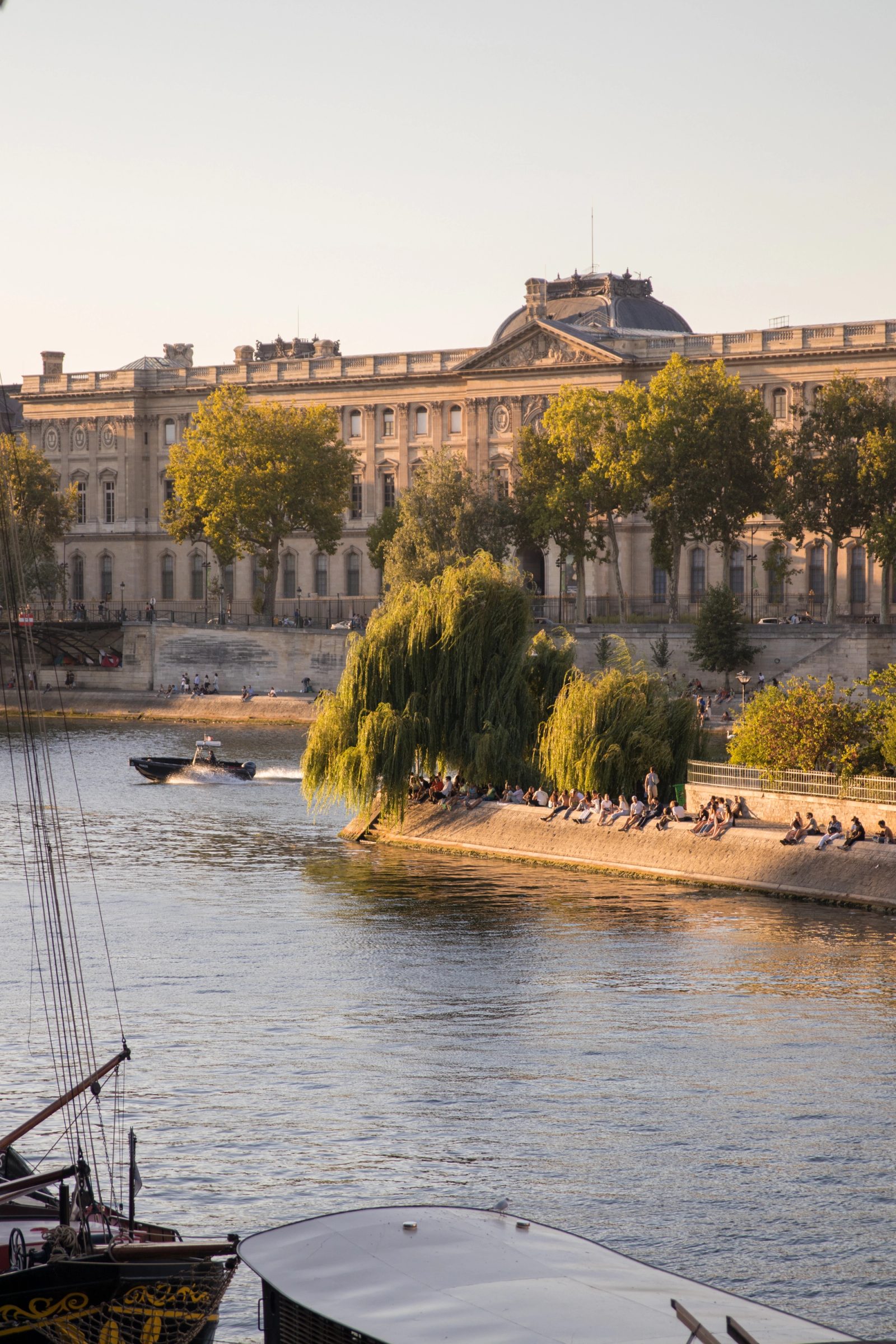 A view across a river showing people sitting on a stone embankment beneath weeping willow trees, with a classical palatial building and boats along the waterfront under soft evening light.