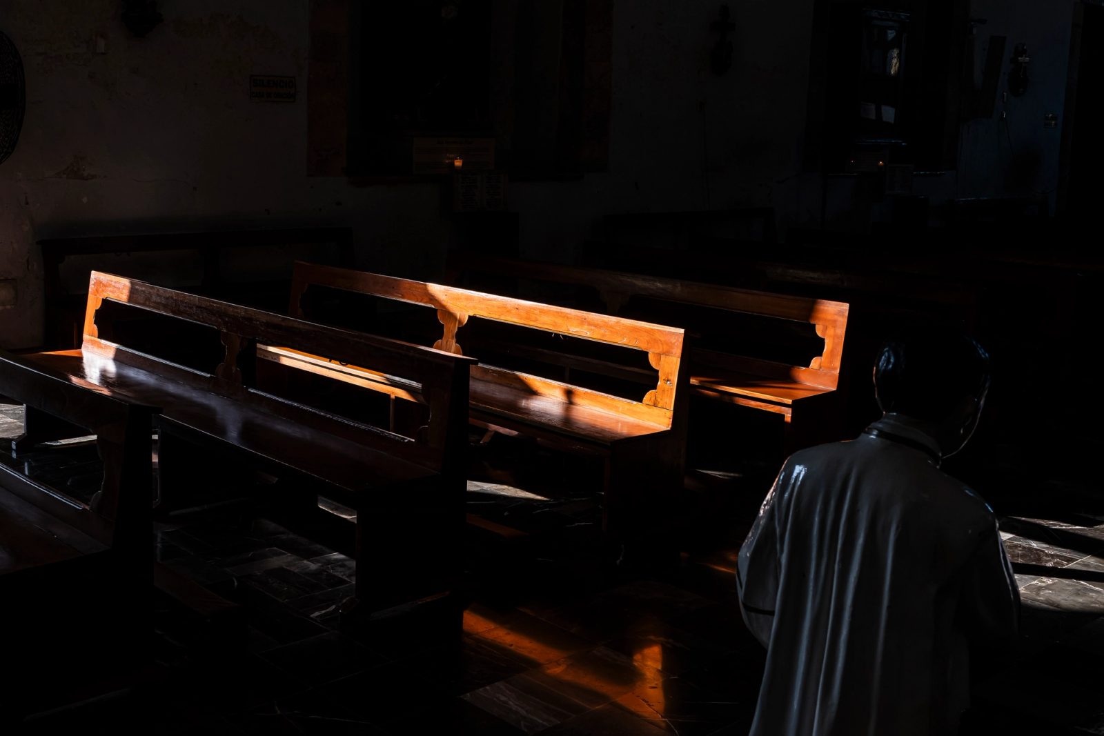 A person in dark clothing kneels beside church pews illuminated by warm sunlight streaming through a window in an otherwise dark interior.