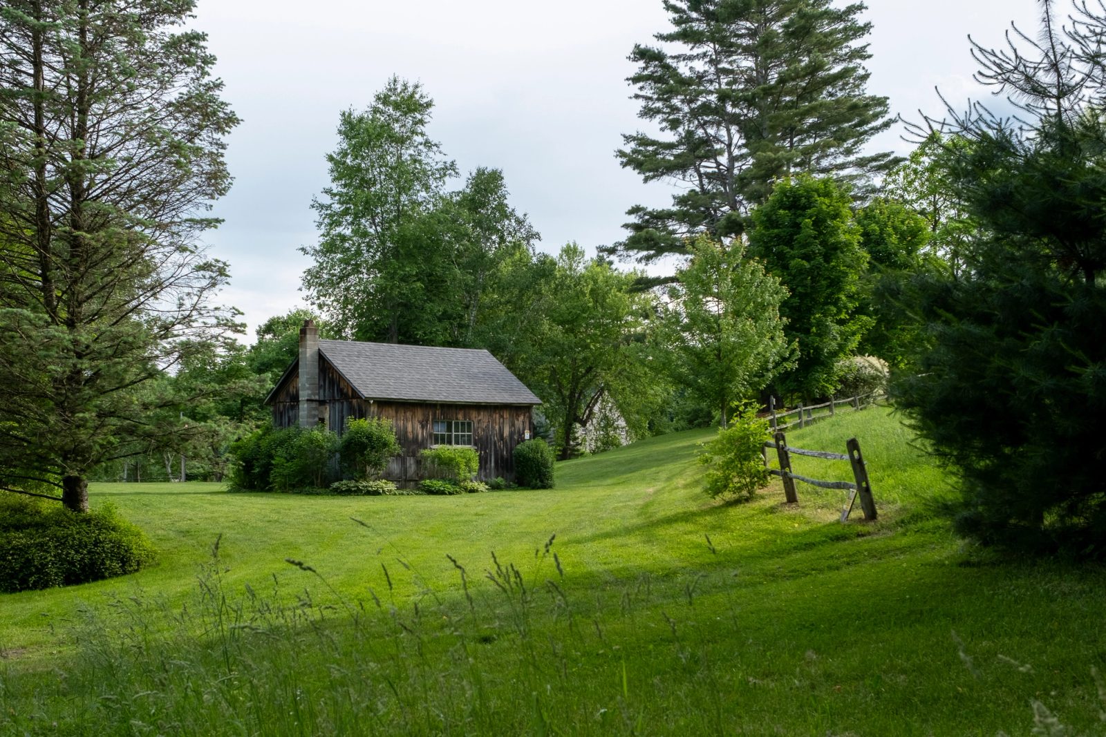 A rustic wooden barn with a gray shingled roof sits on a green hillside surrounded by pine and deciduous trees with an old wooden fence in the foreground.