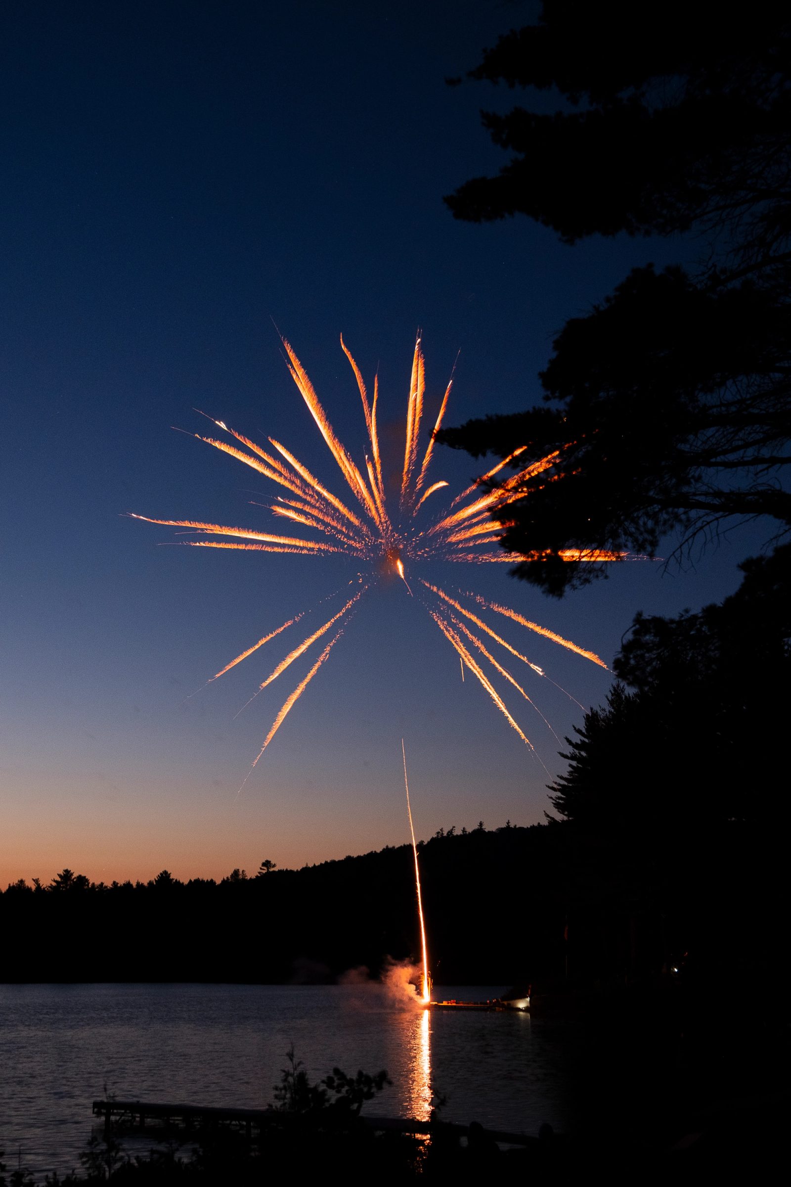 A single orange and gold firework explodes in a starburst pattern over a lake at dusk, with silhouetted trees framing the scene and the firework's reflection visible on the water.