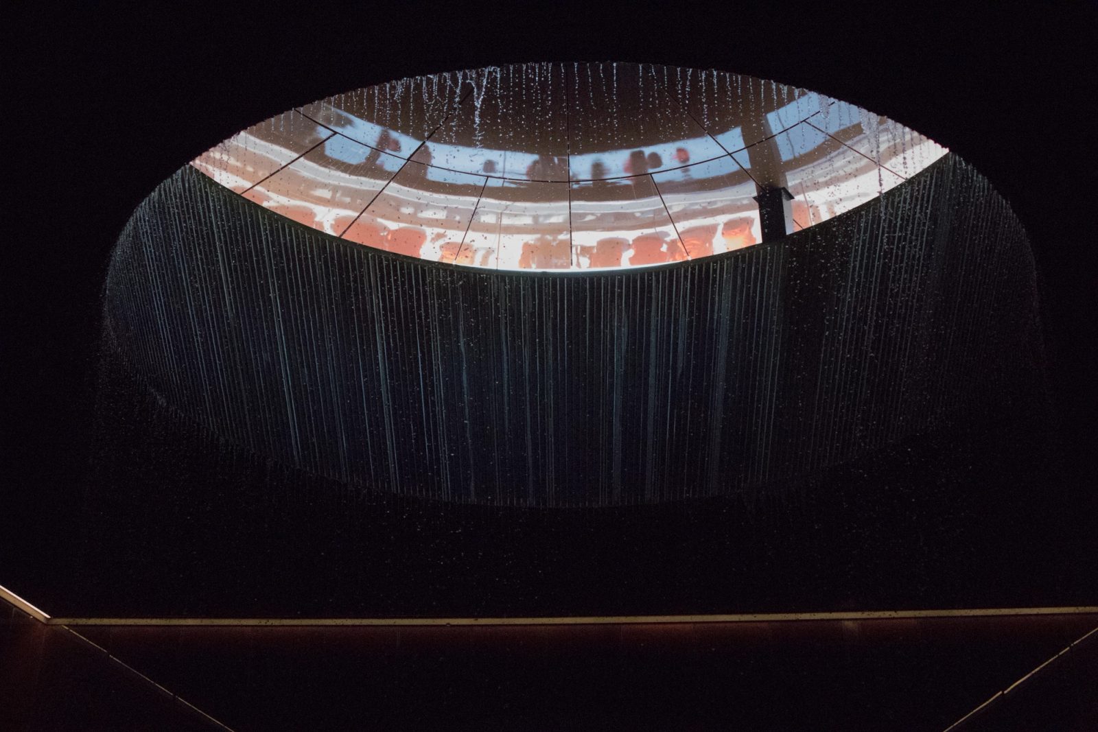 A circular skylight viewed from below shows illuminated water droplets falling in vertical streams around its perimeter, with evening sky and power lines visible through the opening.