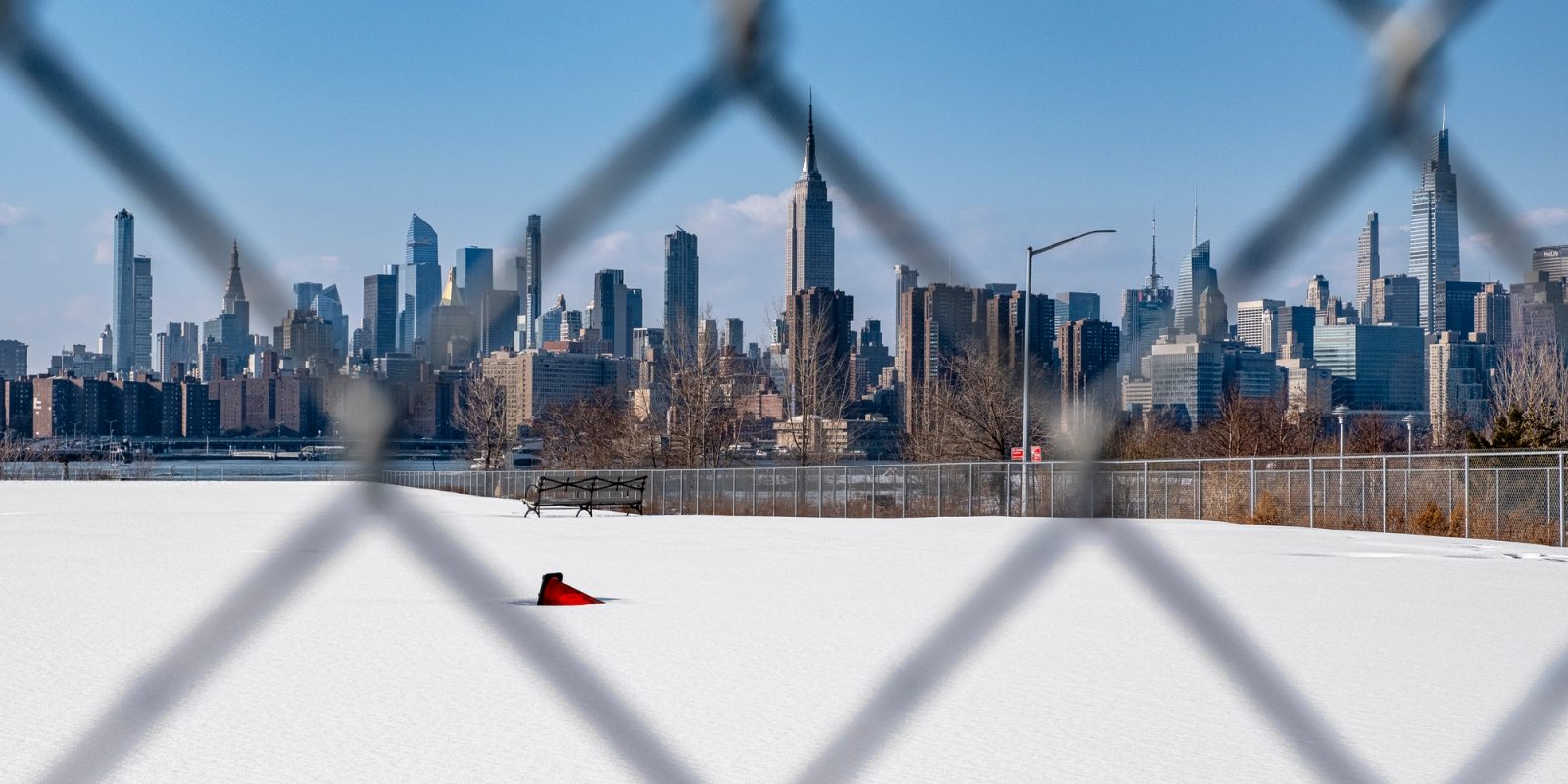 View of a city skyline through a chain-link fence with snow-covered field in the foreground and a red object on the white snow.