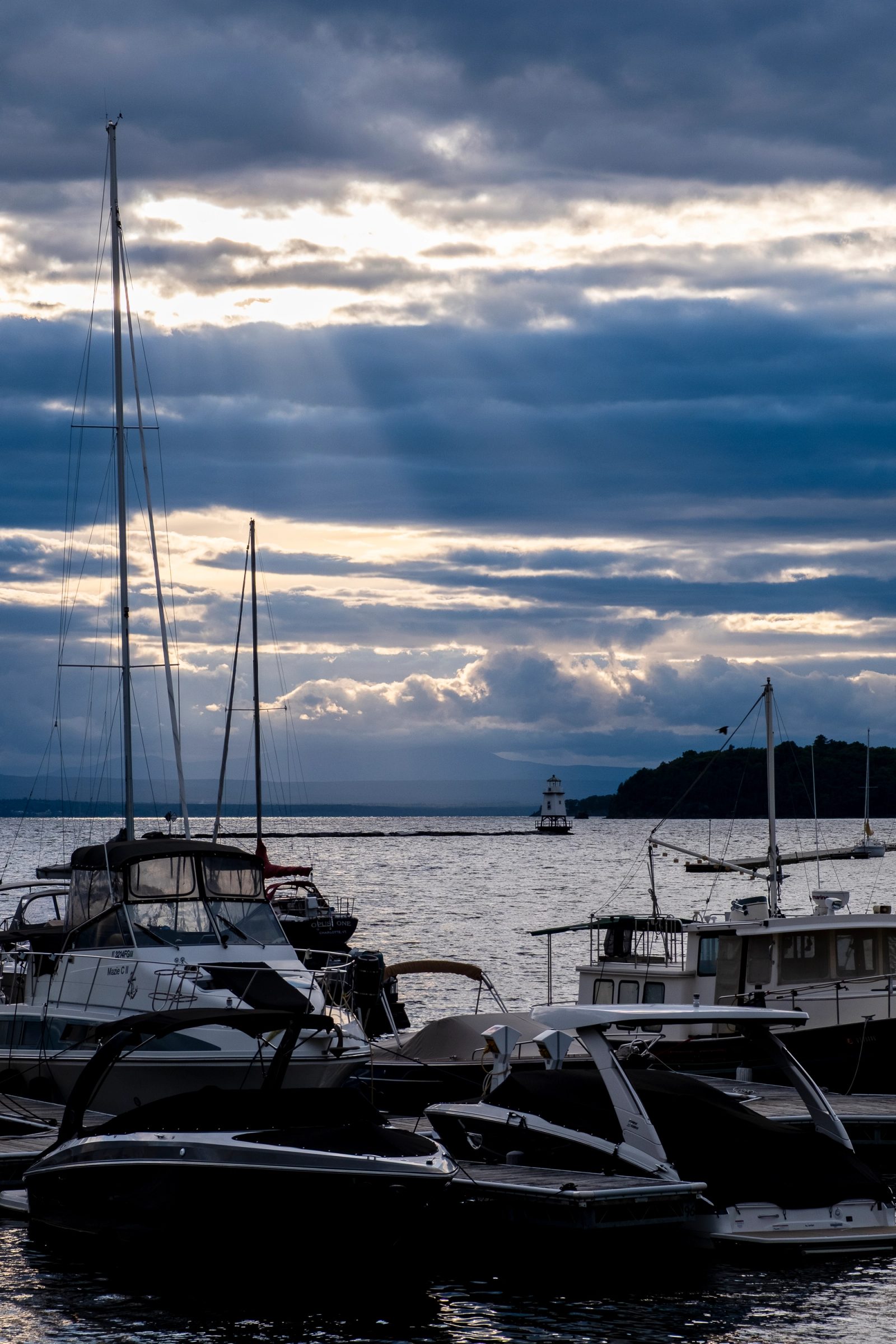 A marina with silhouetted boats in the foreground under dramatic storm clouds with rays of sunlight breaking through over a harbor with a lighthouse visible in the distance.