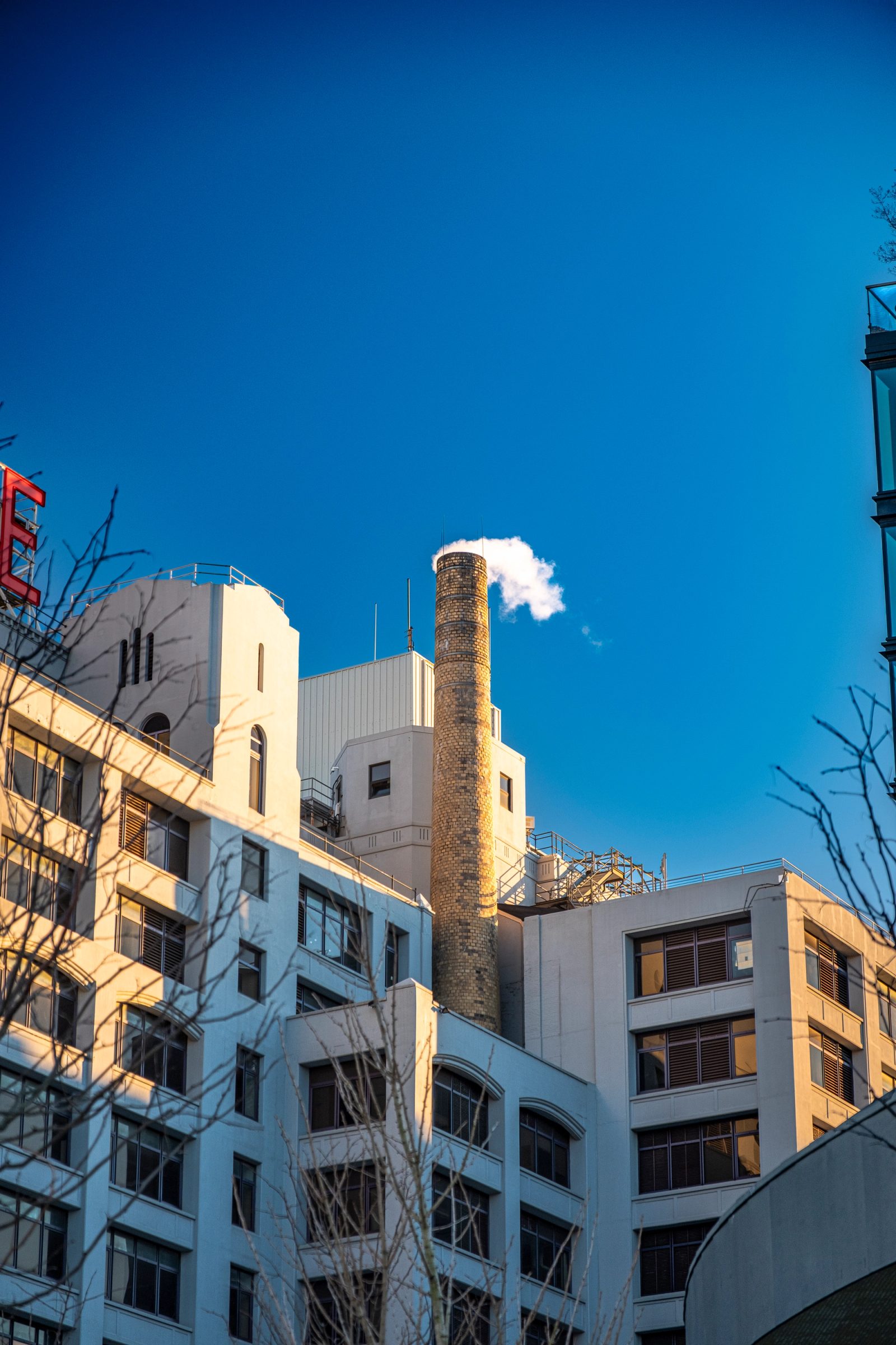 An old brick smokestack emitting white steam stands among white urban buildings under a clear blue sky.