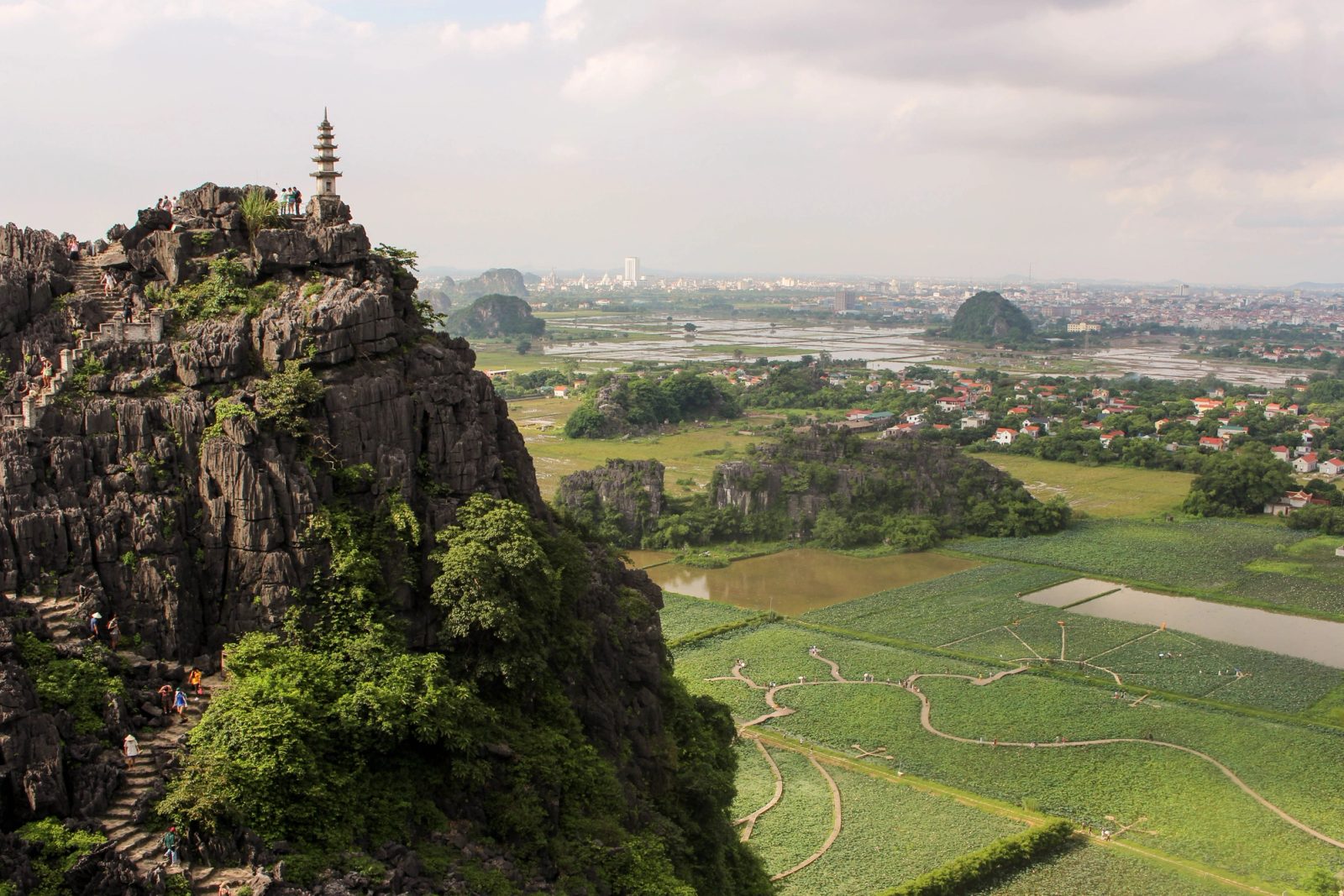 A multi-tiered pagoda sits atop a dramatic rocky limestone outcrop overlooking a valley of rice paddies, with karst formations and a distant city visible under a cloudy sky.