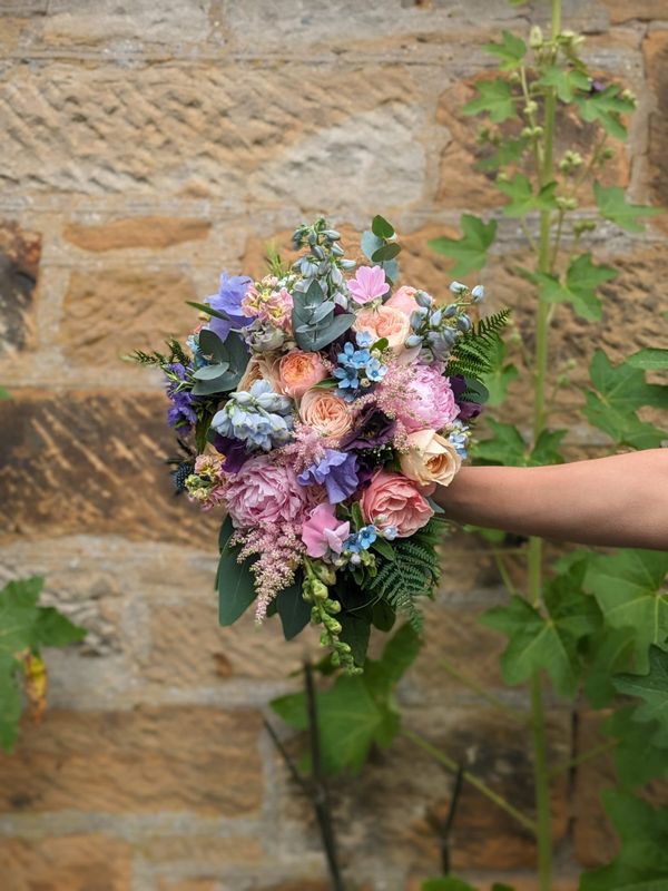 Teardrop-shaped bouquet full of seasonal summer flowers is held for picture in front of stone wall