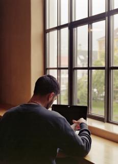 A male student sitting in front of a window and reading.