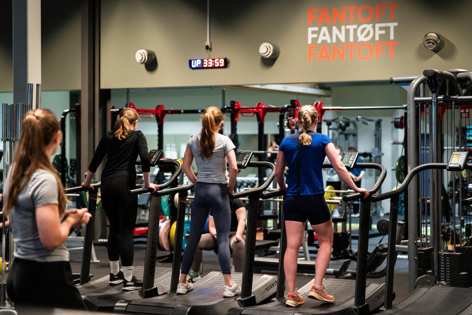 Sett bakfra av flere personer som trener på tredemøller i et treningsstudio med en instruktør som følger med. Det  "FANTOFT - FANTØFT - FANTOFT" skrevet på veggen i bakgrunnen.
