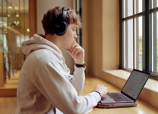 A male student with headphones has a laptop in front of him, reading with concentration.
