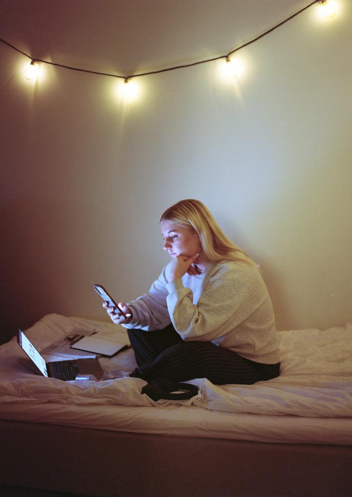 Kvinnelig student som sitter i sengen med pc og mobil.