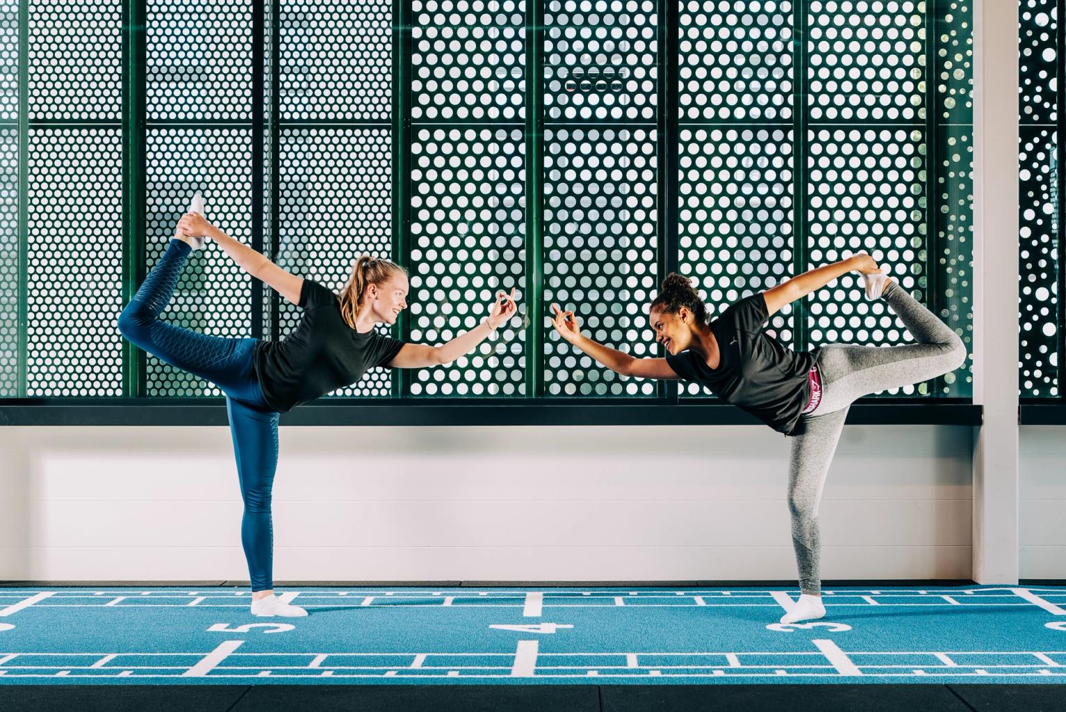  Two women face each other while balancing on one foot. They are at a fitness center, dressed in workout attire.