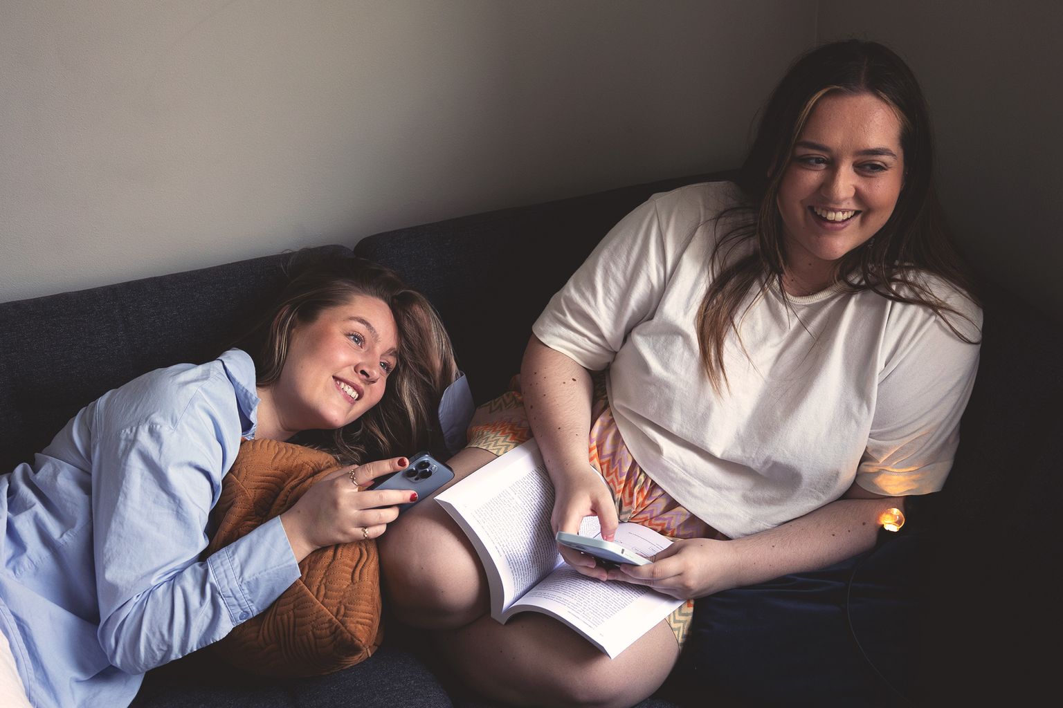 To kvinner sitter sammen med mobiltelefonene sine og en bok, deler smil mens de nyter et øyeblikk fylt med latter.
