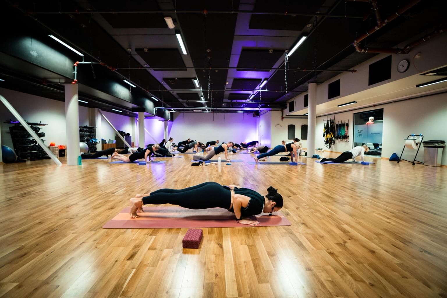A large gym with a parquet floor and mirrored walls, where a group of people are doing a yoga exercise on exercise mats during a group session.