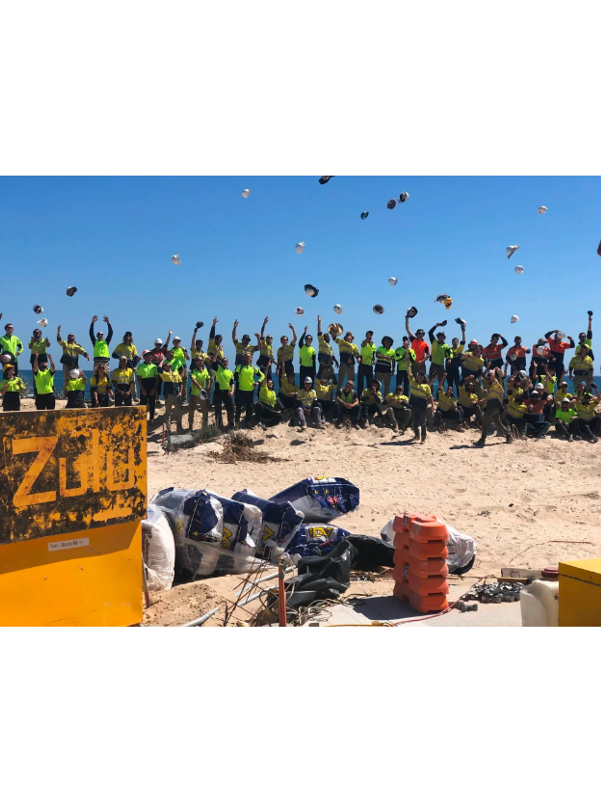 Large group of people in safety vests and helmets, celebrating with raised arms at a team-building event on a sandy site