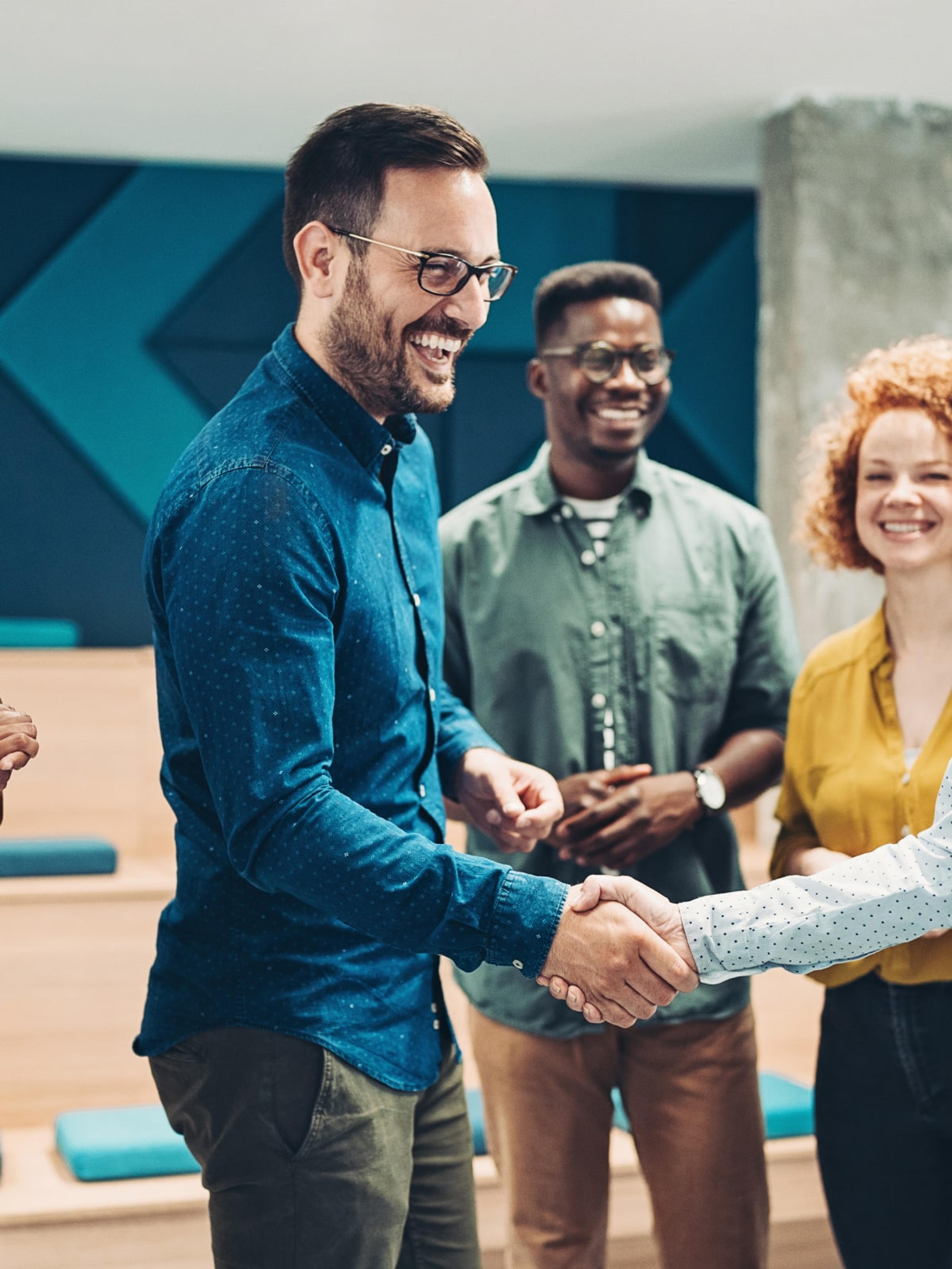A business coach shaking hands with a team member in a modern office environment, surrounded by a diverse group of colleagues, illustrating the collaborative and supportive nature of Adapt's coach-guided program.