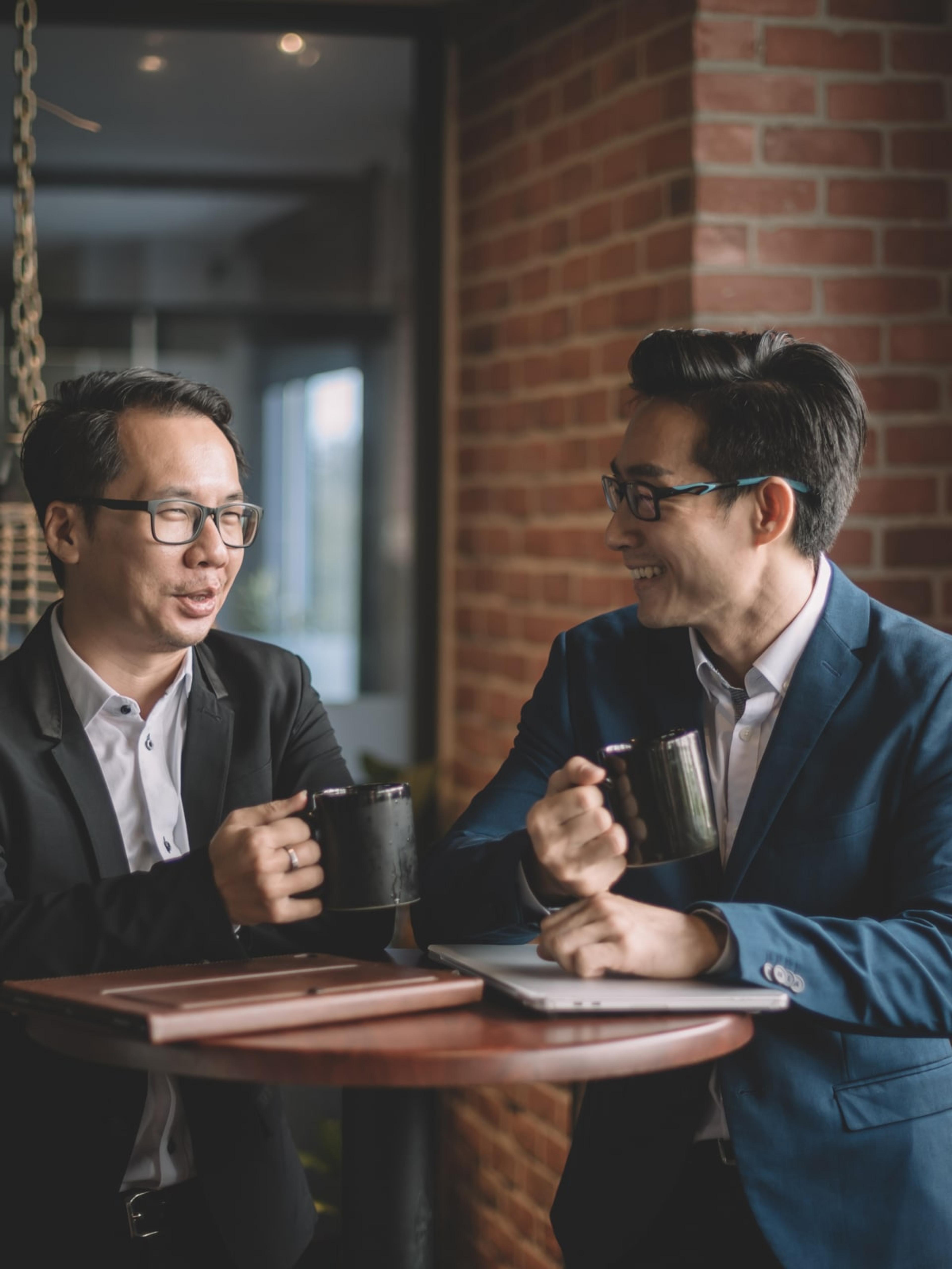 Two business professionals in suits having a casual conversation over coffee, illustrating the supportive and collaborative environment fostered by Adapt's coach-guided services.