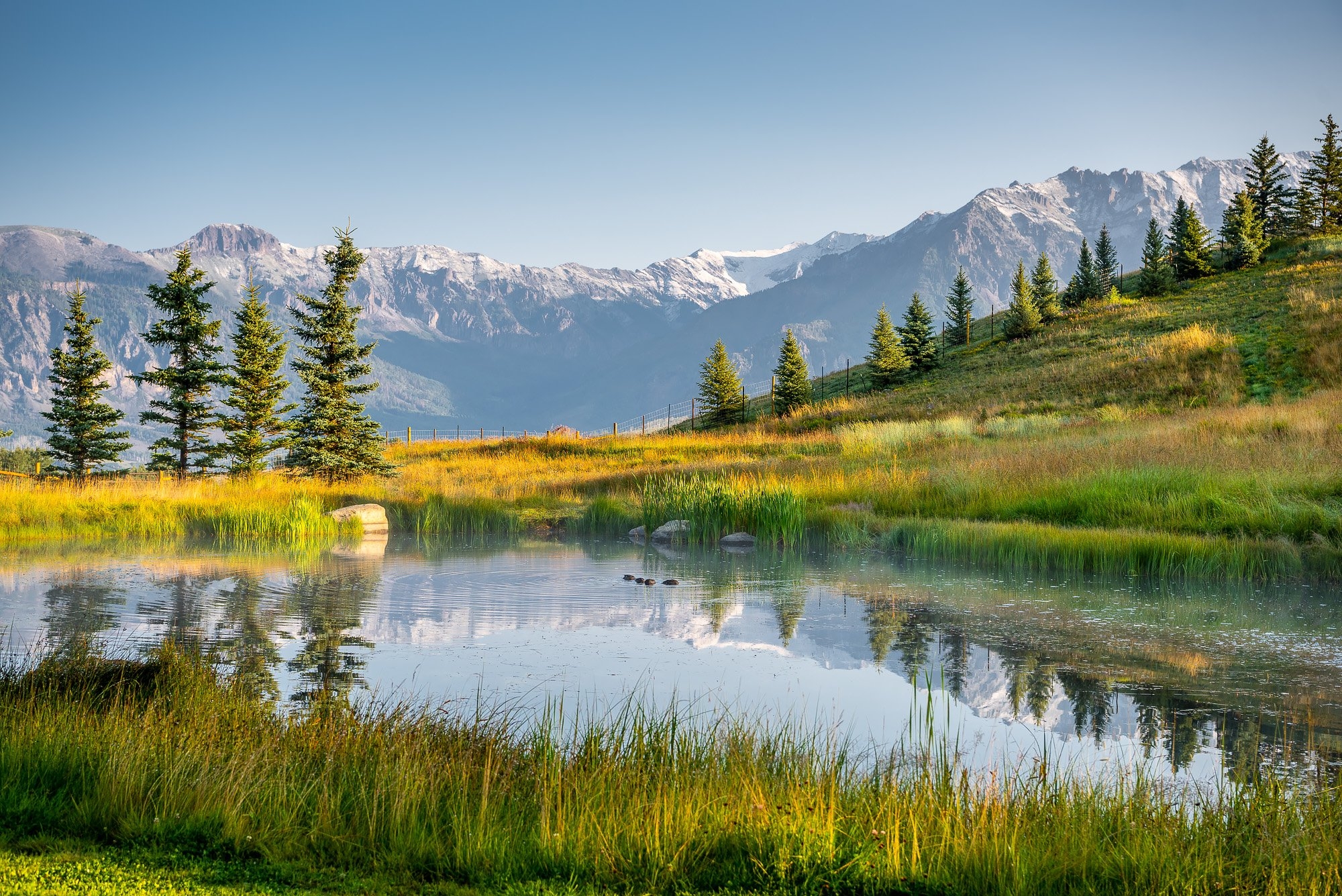 A painting of a pond surrounded by grassy hills near a mountain