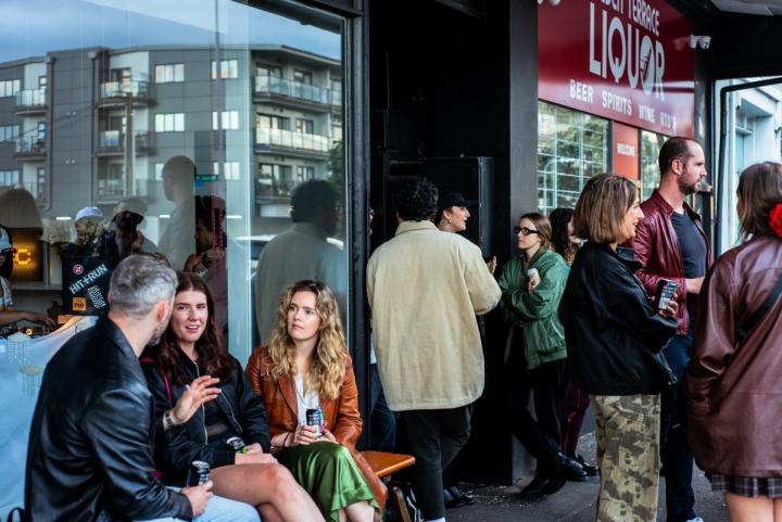 Community members gathered in conversation outside TBC Radio while the DJ can be seen through the window playing vinyl.