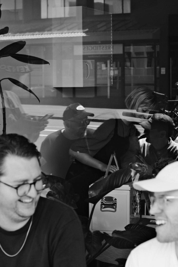  Community members laughing on a bench in front of the space. You can see reflections in the window of more community members, as well as market vendors inside through the window.