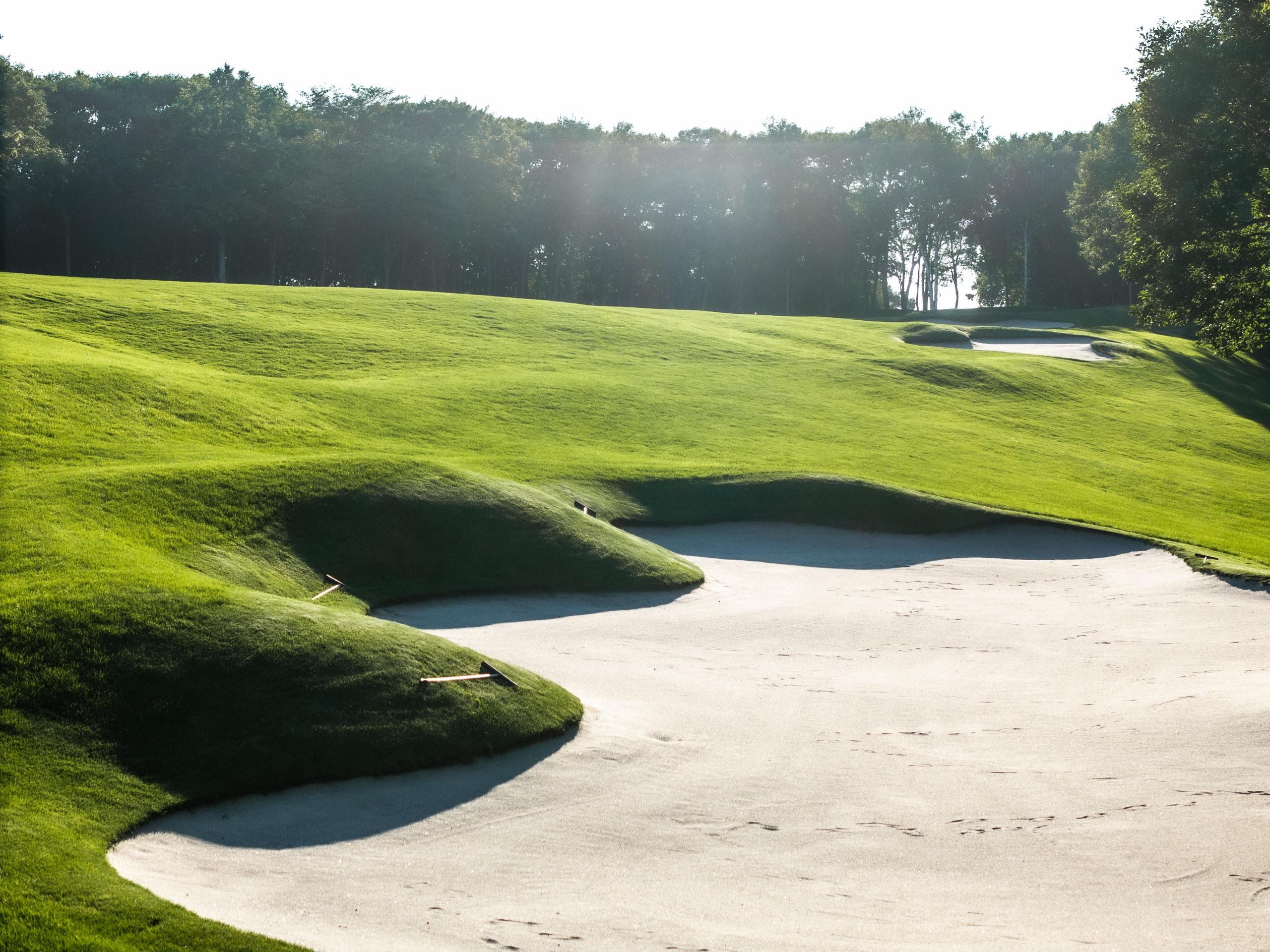 Deep greenside bunker set into grass-covered mounds in low evening sunlight