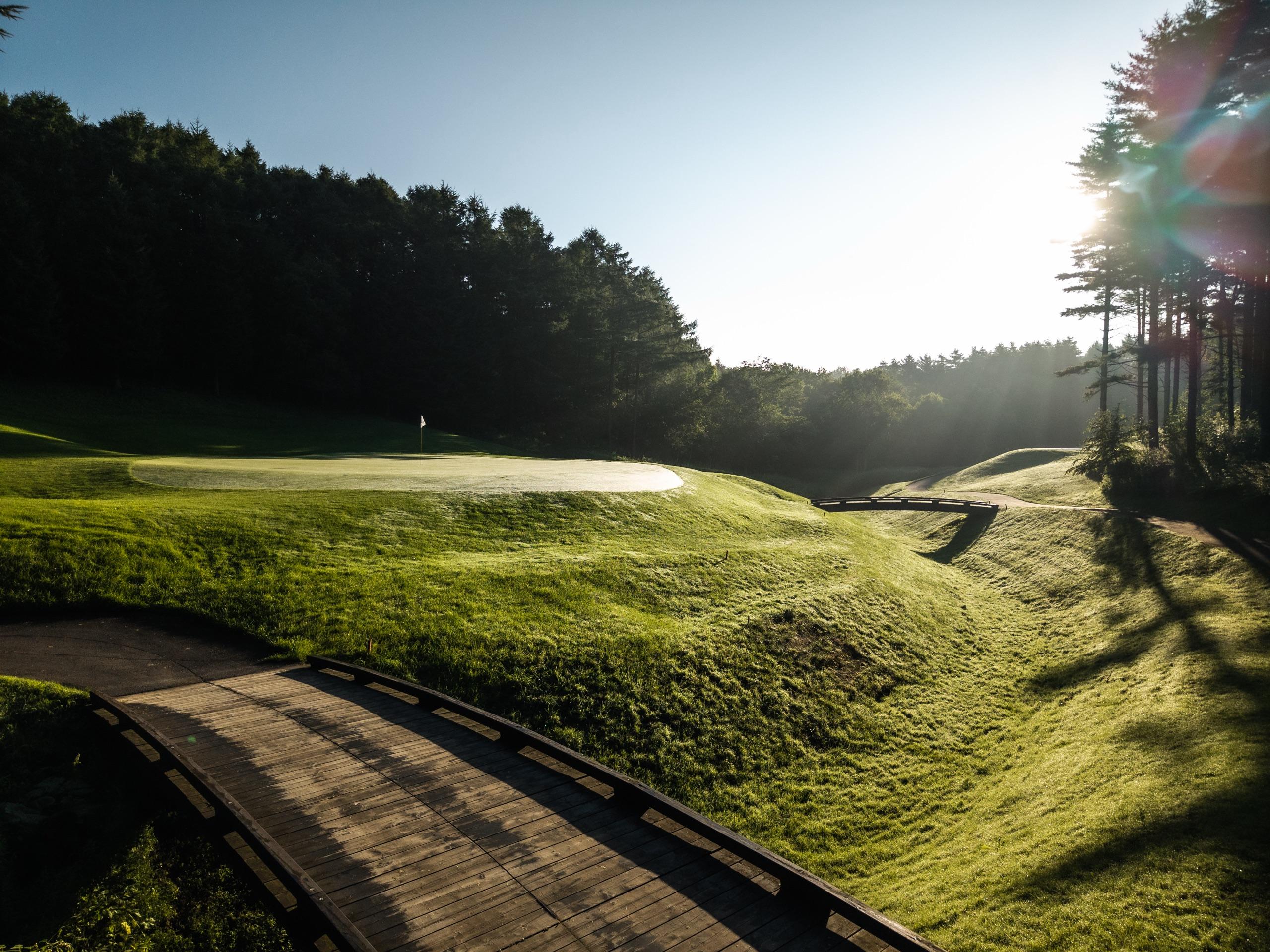 Wooden cart bridge leading toward a morning-lit green set against pine forest at Katsura Golf Club