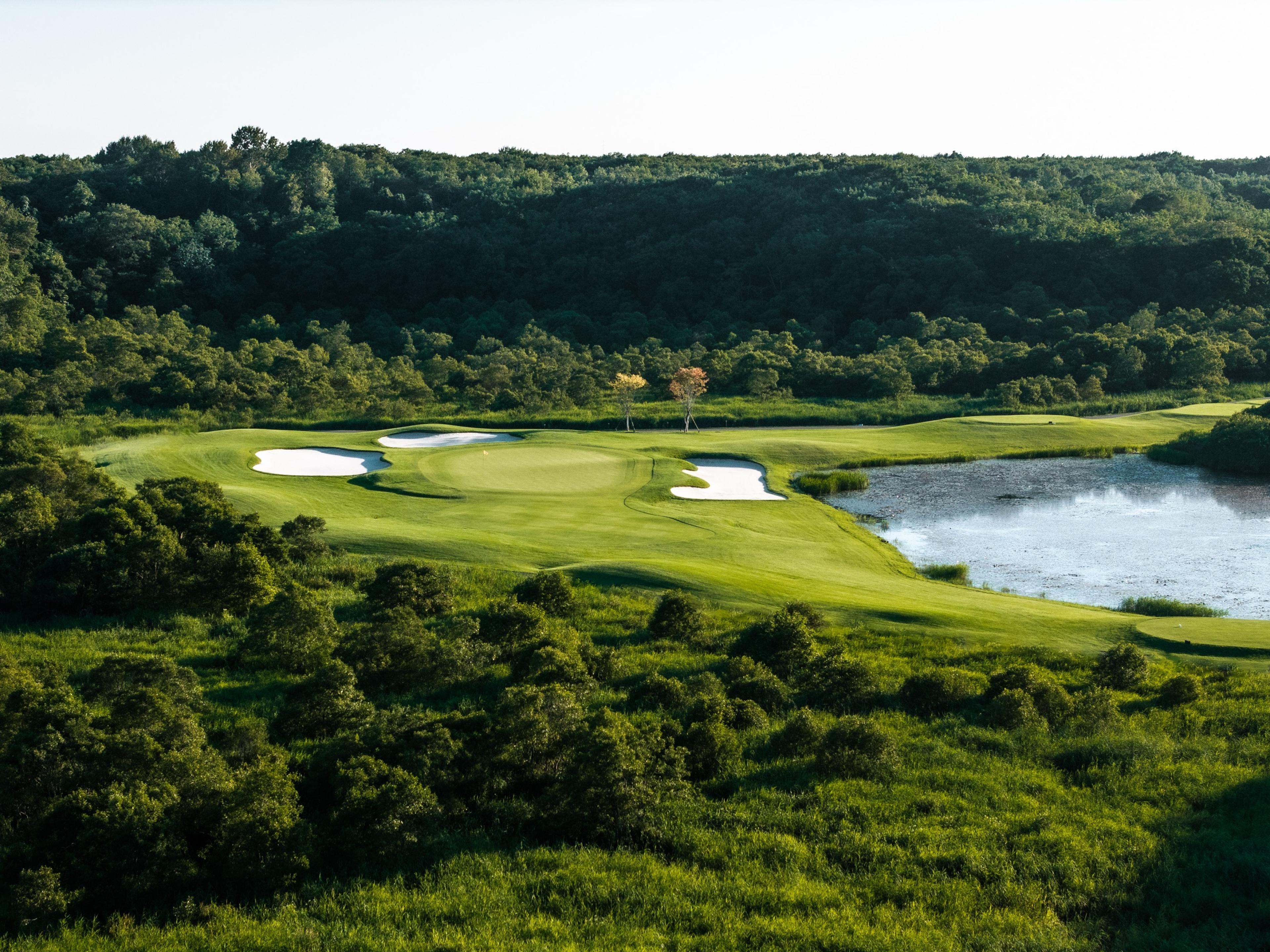 Elevated view over a green complex ringed by white bunkers beside a pond, framed by unbroken forest