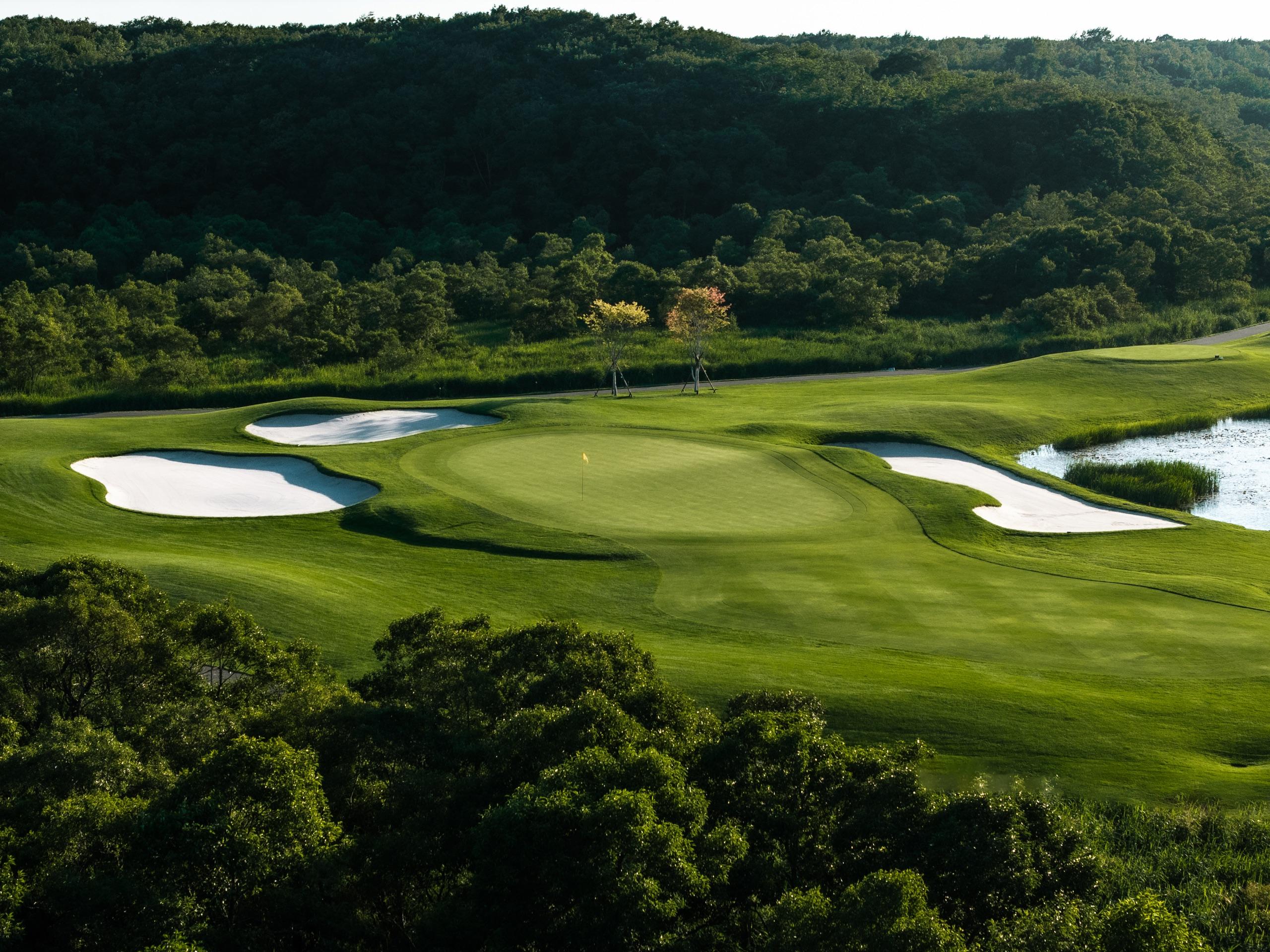 Green defended by four bunkers at the water's edge, backed by wooded hills in evening light
